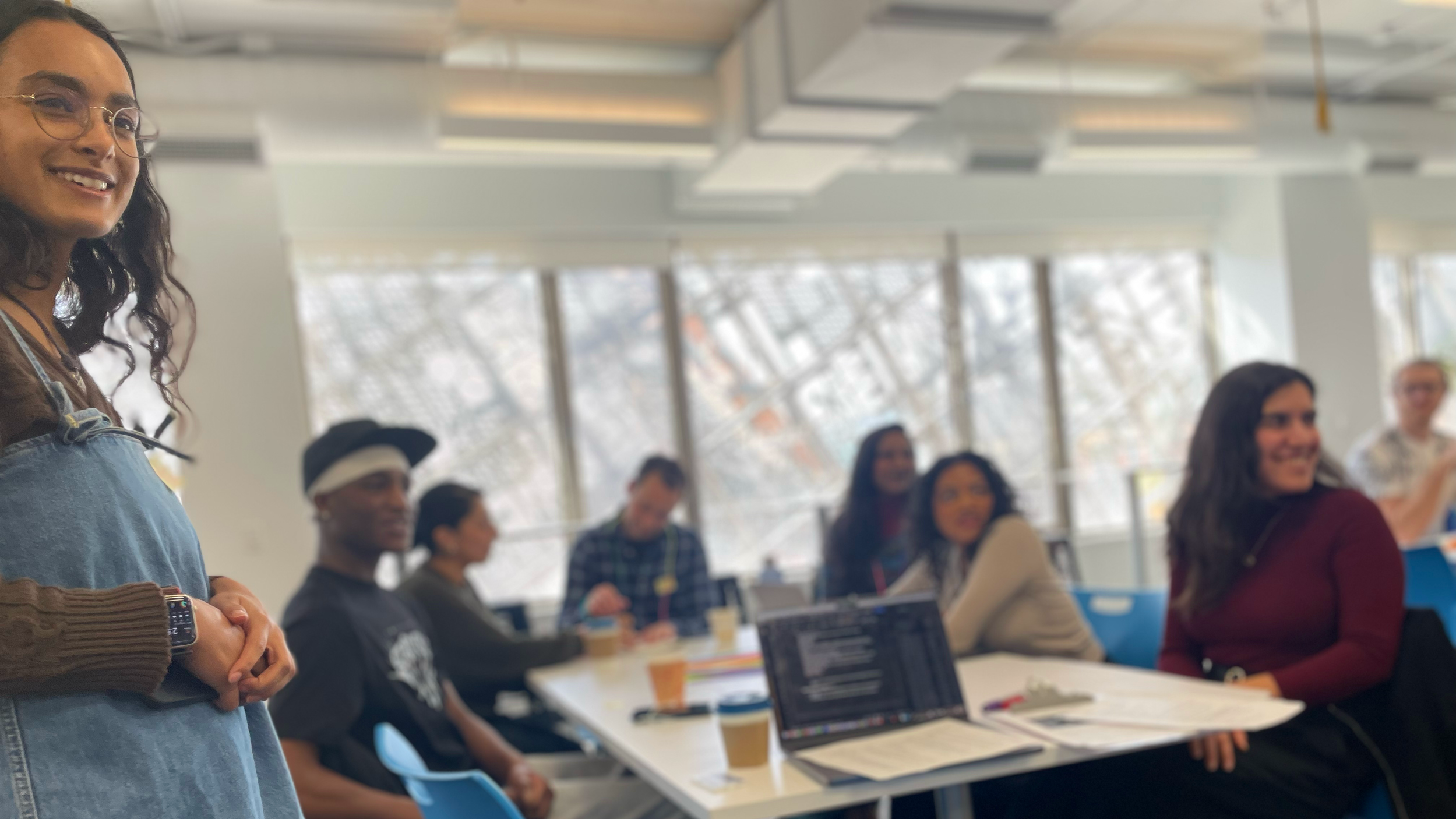 A group of diverse women seated at a table in a bright, modern conference room with large windows. One woman stands at the front smiling, wearing glasses, a brown sweater, and a denim apron, possibly leading a discussion or presentation.