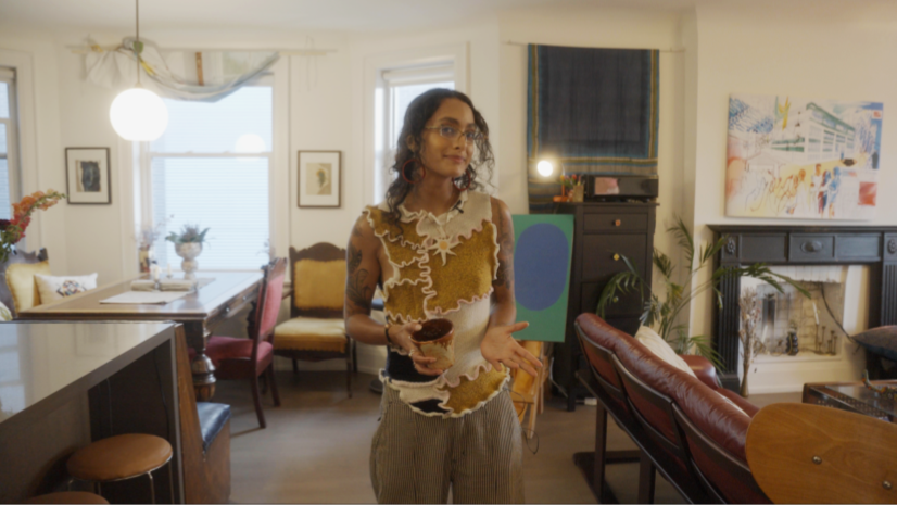 A woman with glasses and tattoos standing in a well-decorated living room, holding a bowl in her left hand, with her right hand gesturing. The room has a dining area with a wooden table and pink chairs, a window with sheer curtains, artwork on the wa