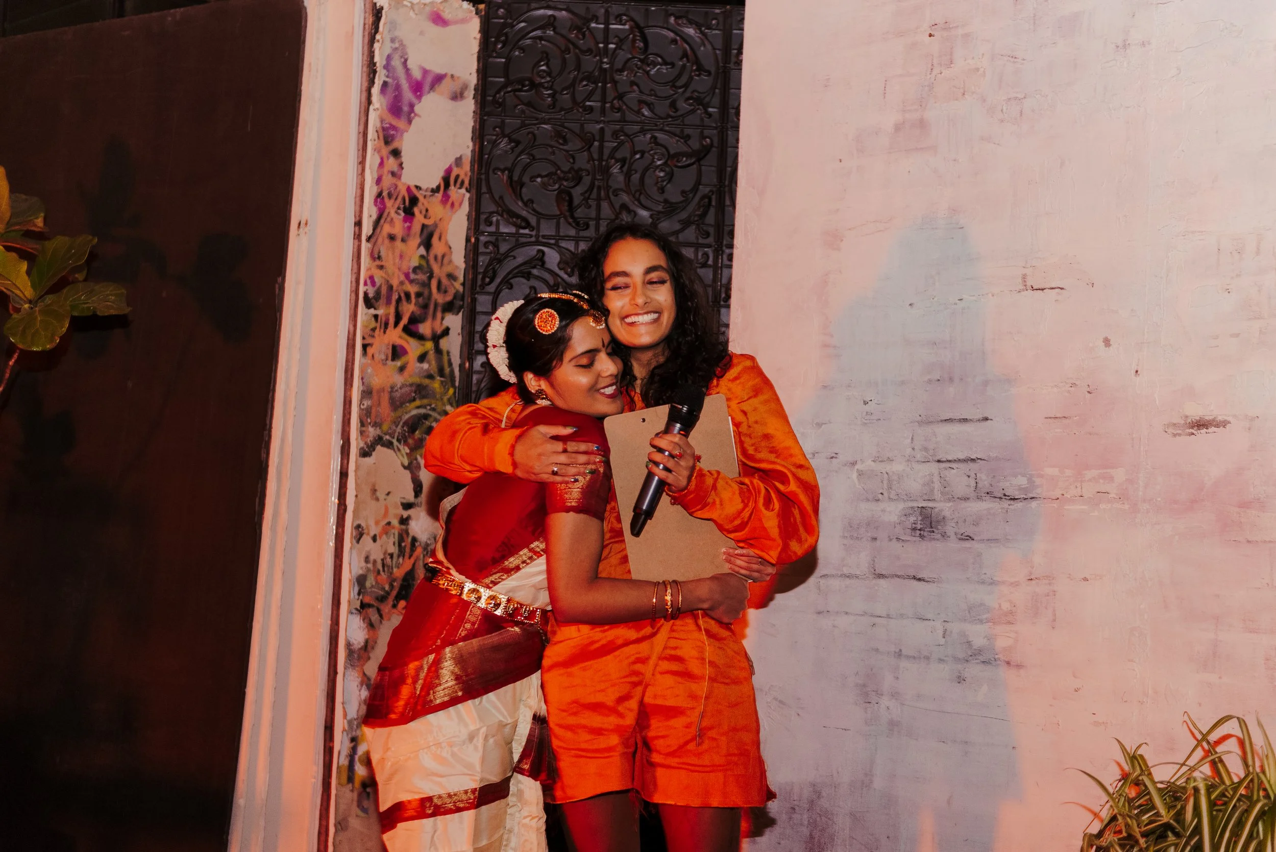 Two women are hugging each other warmly, one holding a microphone and a clipboard, both smiling happily. They are dressed in colorful traditional and modern outfits, standing against a wall with artistic decorations.