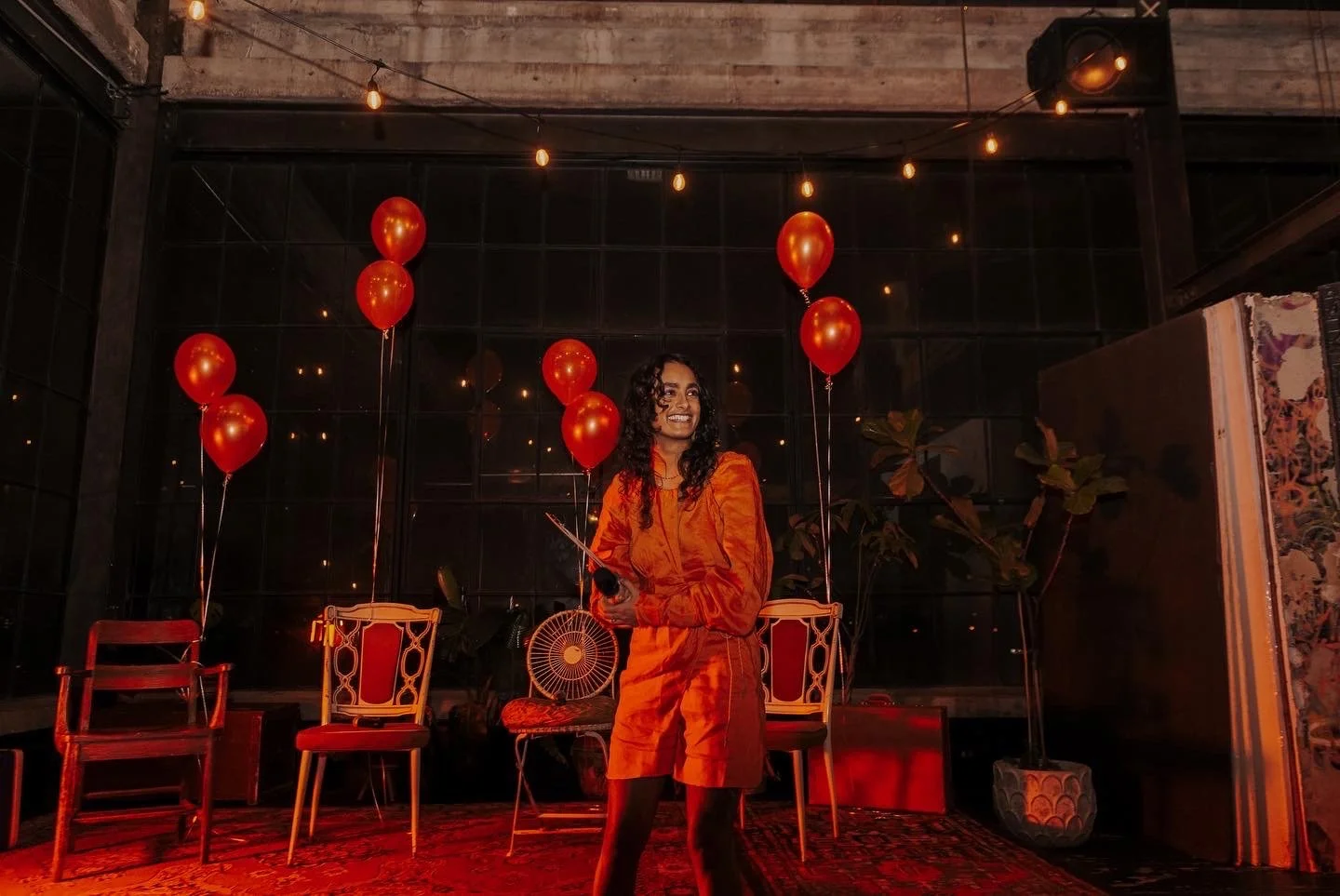 A woman in an orange dress smiling on stage, surrounded by red balloons and vintage chairs, in a dimly lit room with string lights.