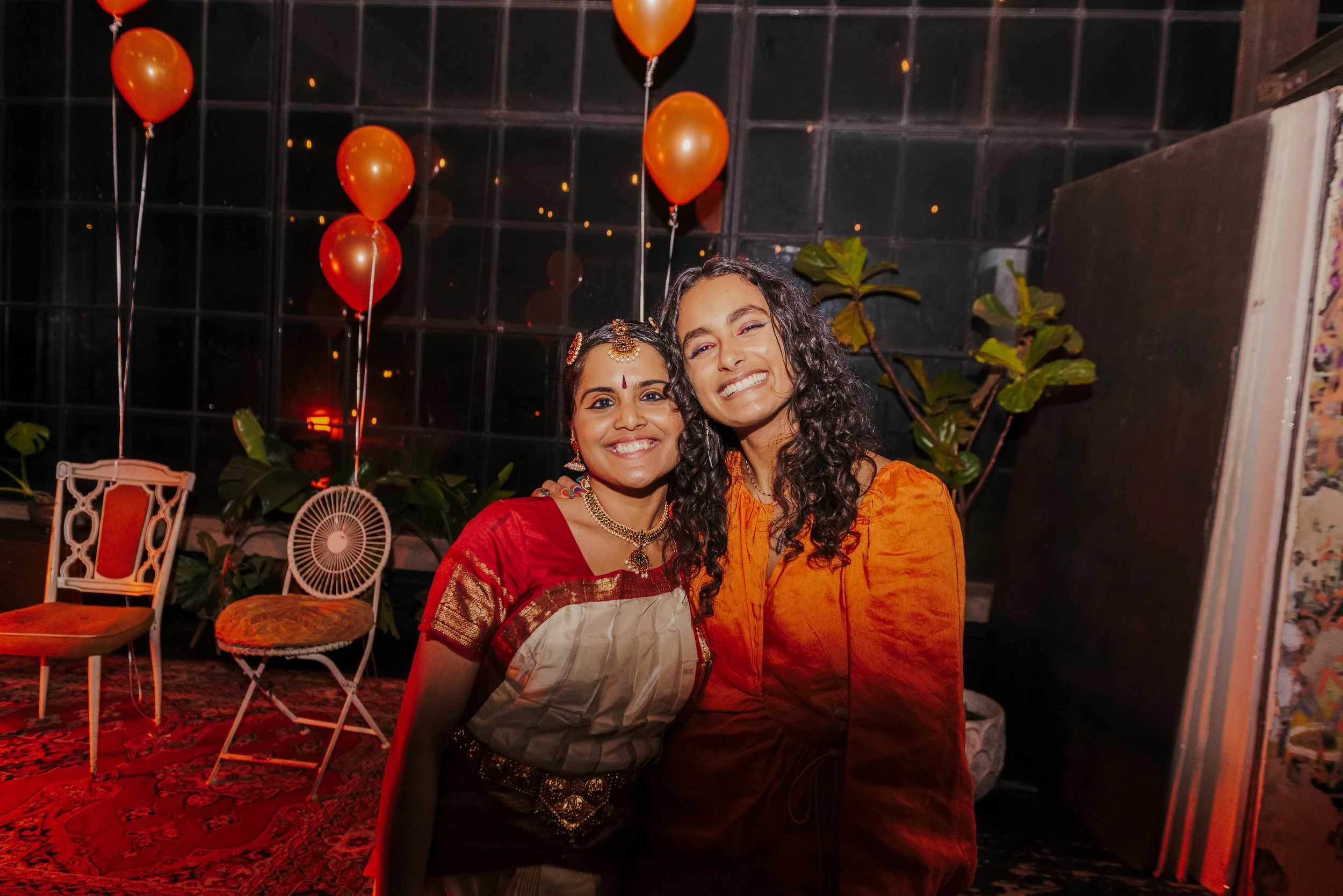 Two women smiling at a celebration or party, with red balloons in the background, sitting on an ornate carpet surrounded by chairs and plants.
