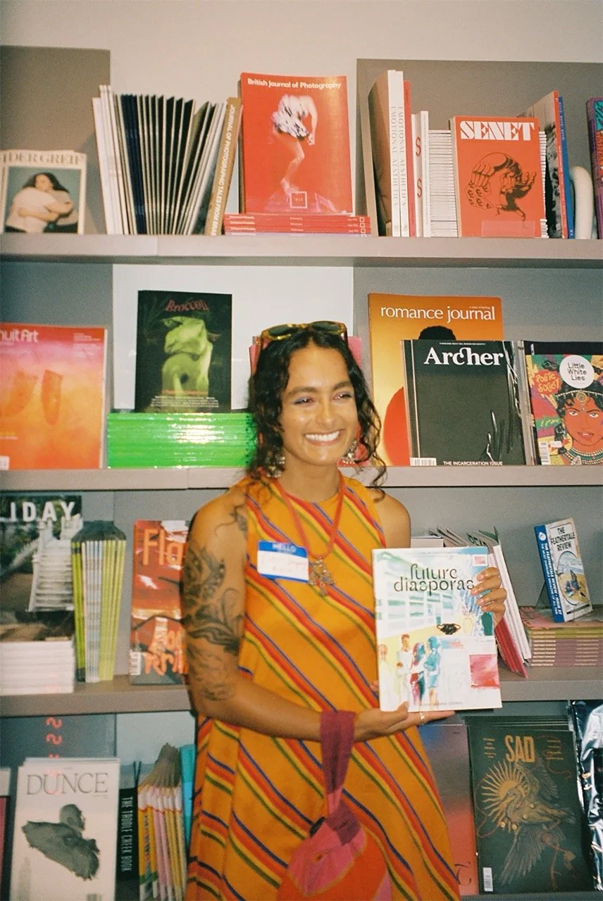A woman with curly dark hair, wearing a colorful striped dress, stands in front of bookshelves at a bookstore, holding a book titled 'Future Diasporas.'