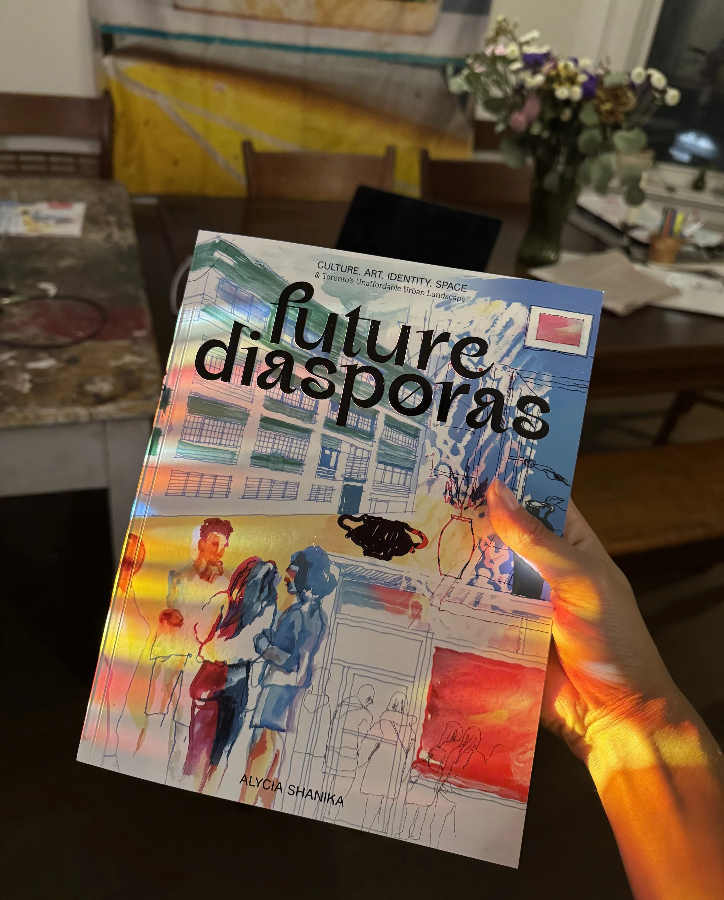 A person holds a book titled 'Future Diasporas' by Alycia Shanika, with a colorful, watercolor-style cover in a room with a wooden table, chairs, a bouquet of flowers, and artwork on the walls.