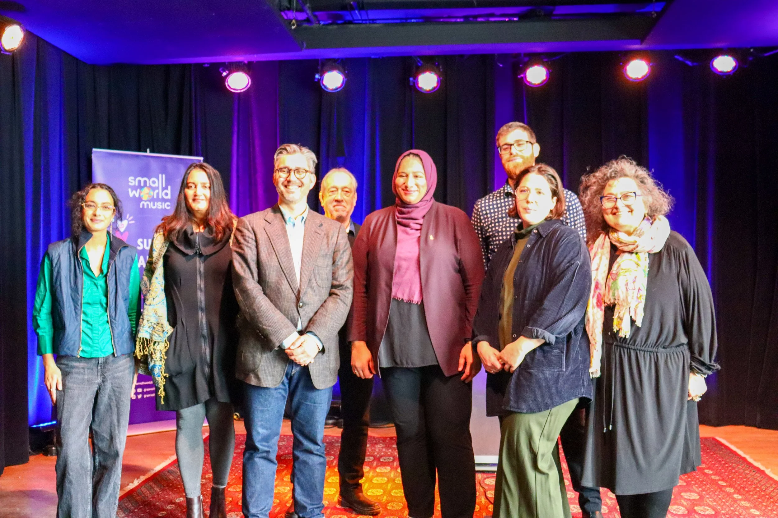 Group of nine people standing on stage with purple lighting, black curtains, and a small world music banner in the background.