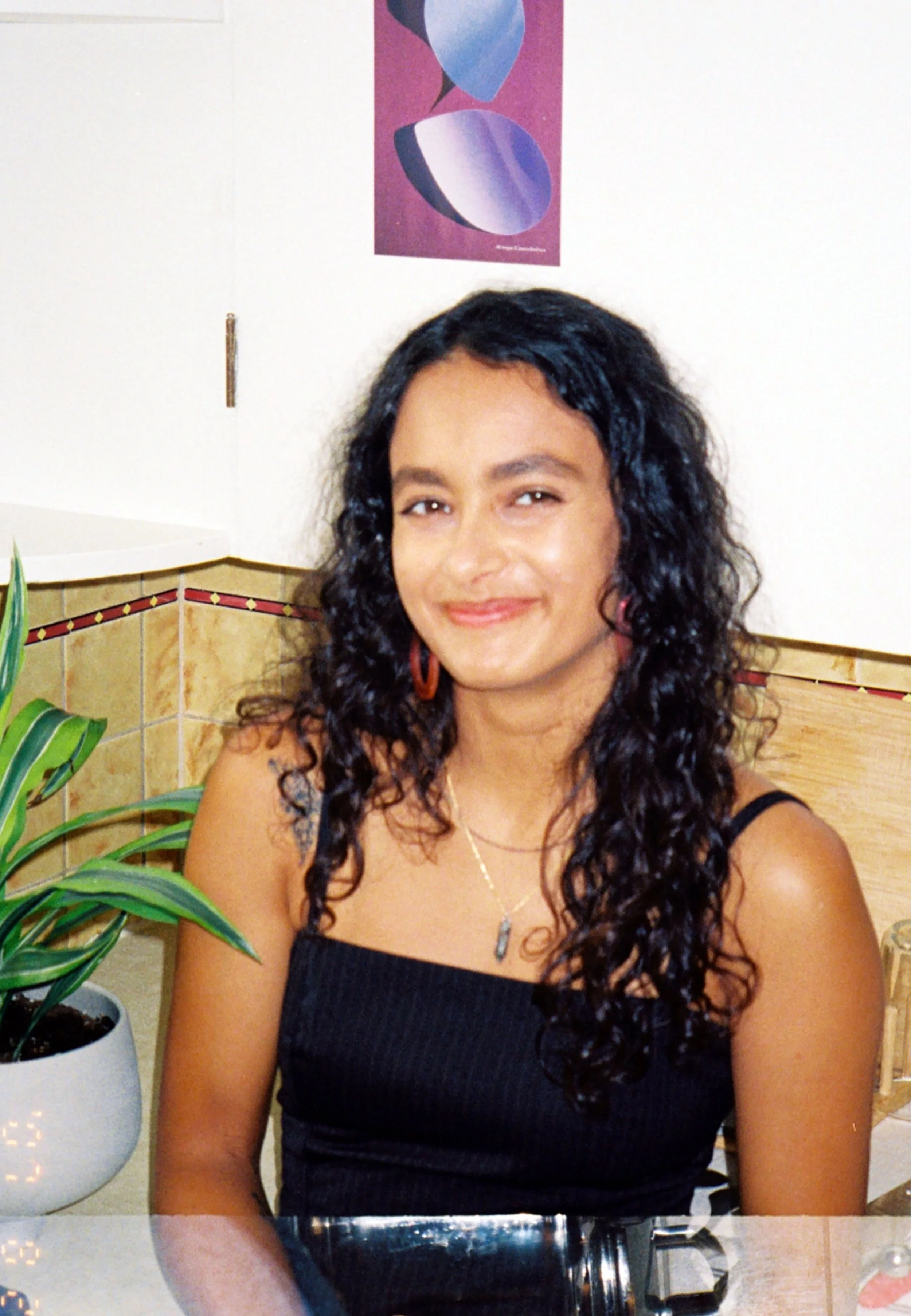 A woman with long curly black hair, wearing a black sleeveless top, sitting at a table in a restaurant or cafe. She is smiling and looking at the camera, with a potted plant and a small decorative object on the table. The background includes a white 