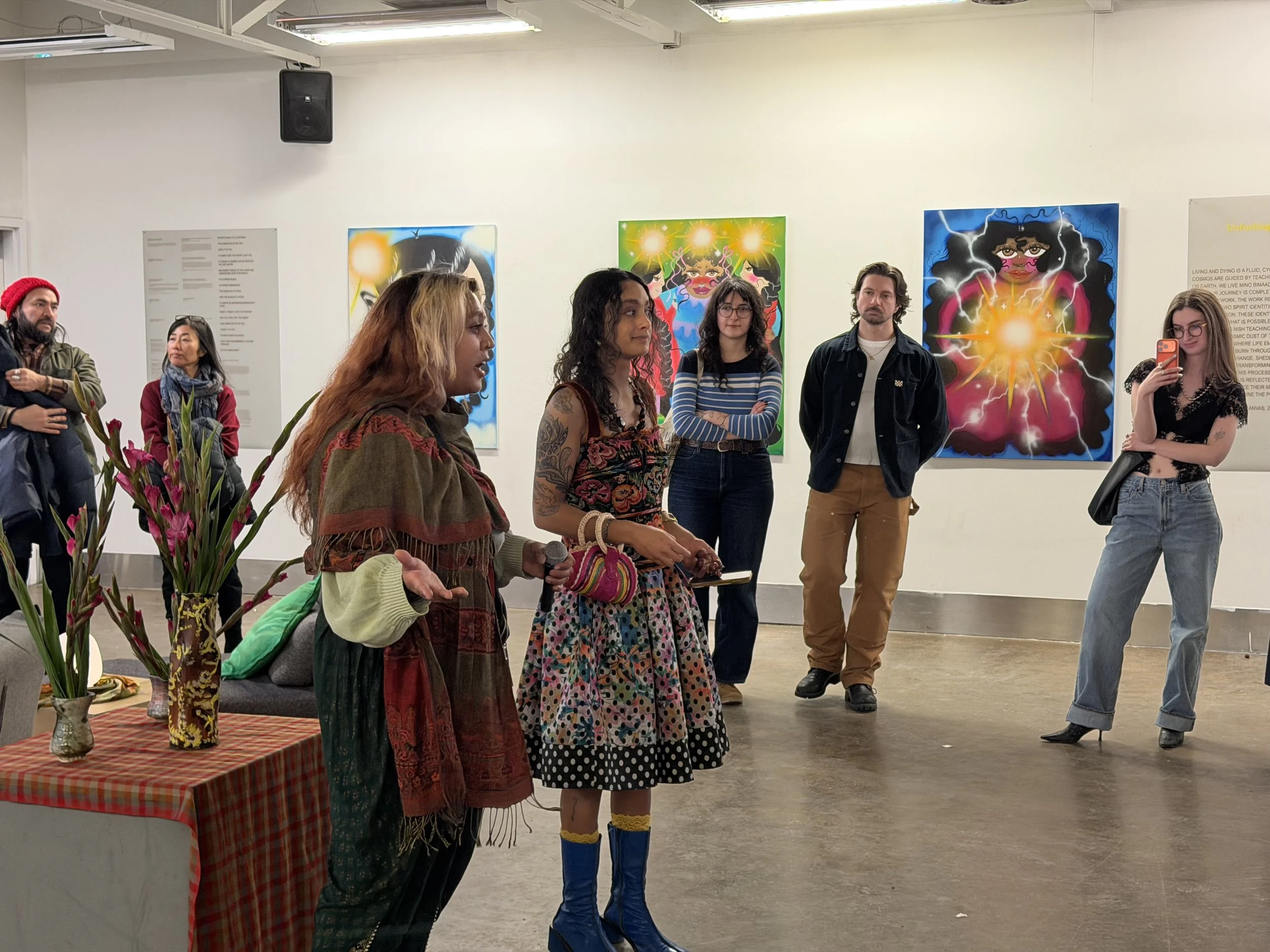 People attending an art exhibit, standing in front of colorful paintings with vibrant and abstract designs. One woman is speaking to the group, holding a microphone, with a table of vases and flowers nearby. Others are listening or taking photos.