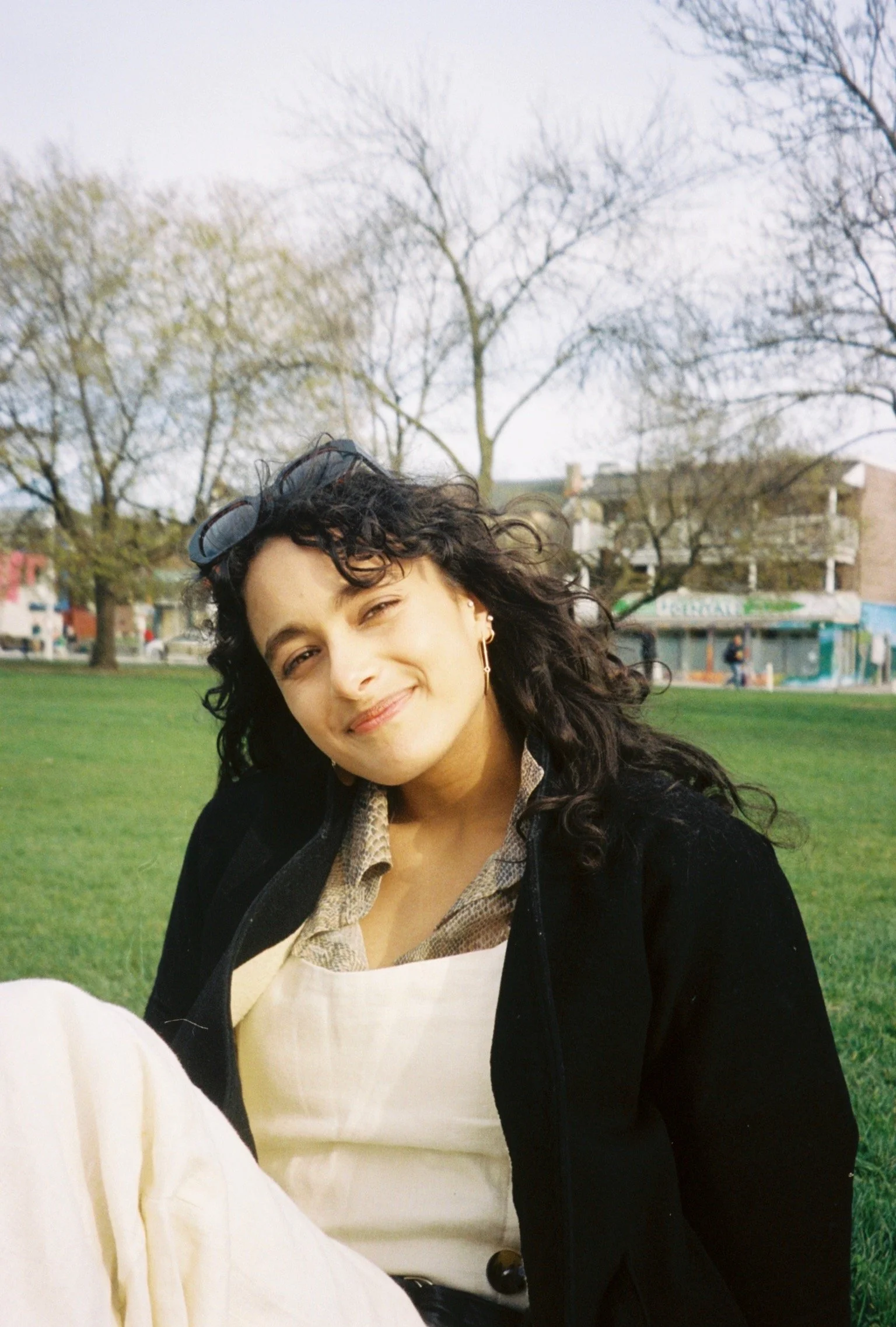 Young woman with curly dark hair, sunglasses on her head, smiling and winking at the camera, sitting on a grassy park with trees and buildings in the background.