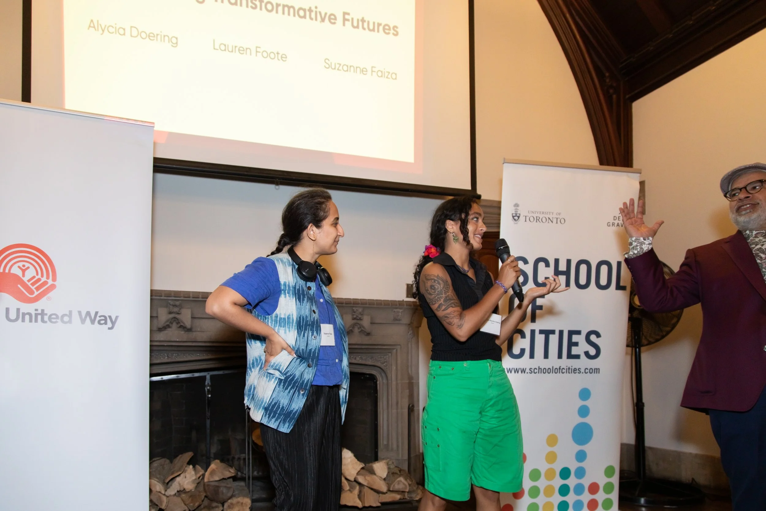 Three people standing on a stage in front of a screen and banners. One woman is speaking into a microphone, another woman is looking at her, and a man is raising his hand. The screen displays names of speakers. One banner is from the United Way, and 