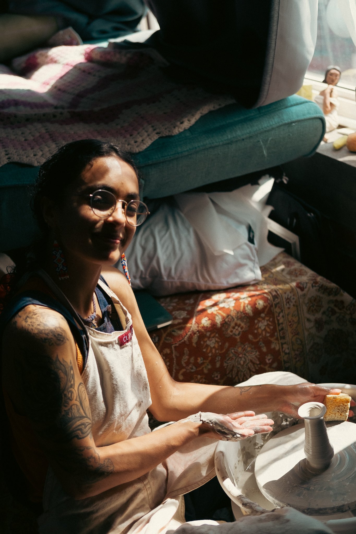 A woman with glasses and tattoos on her arms, wearing earrings and a white apron, is sitting on the floor in a cluttered room. She is smiling at the camera, with some pottery and a sponge in her hands, and a pottery wheel in front of her. There is a 