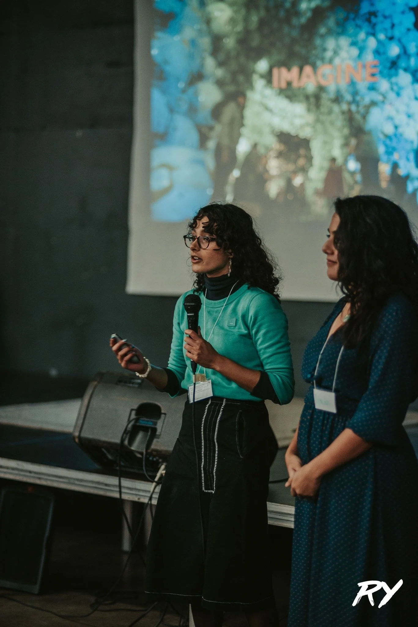 Two women speaking at a conference with a large screen behind them that displays the word 'IMAGINE' and colorful abstract artwork.