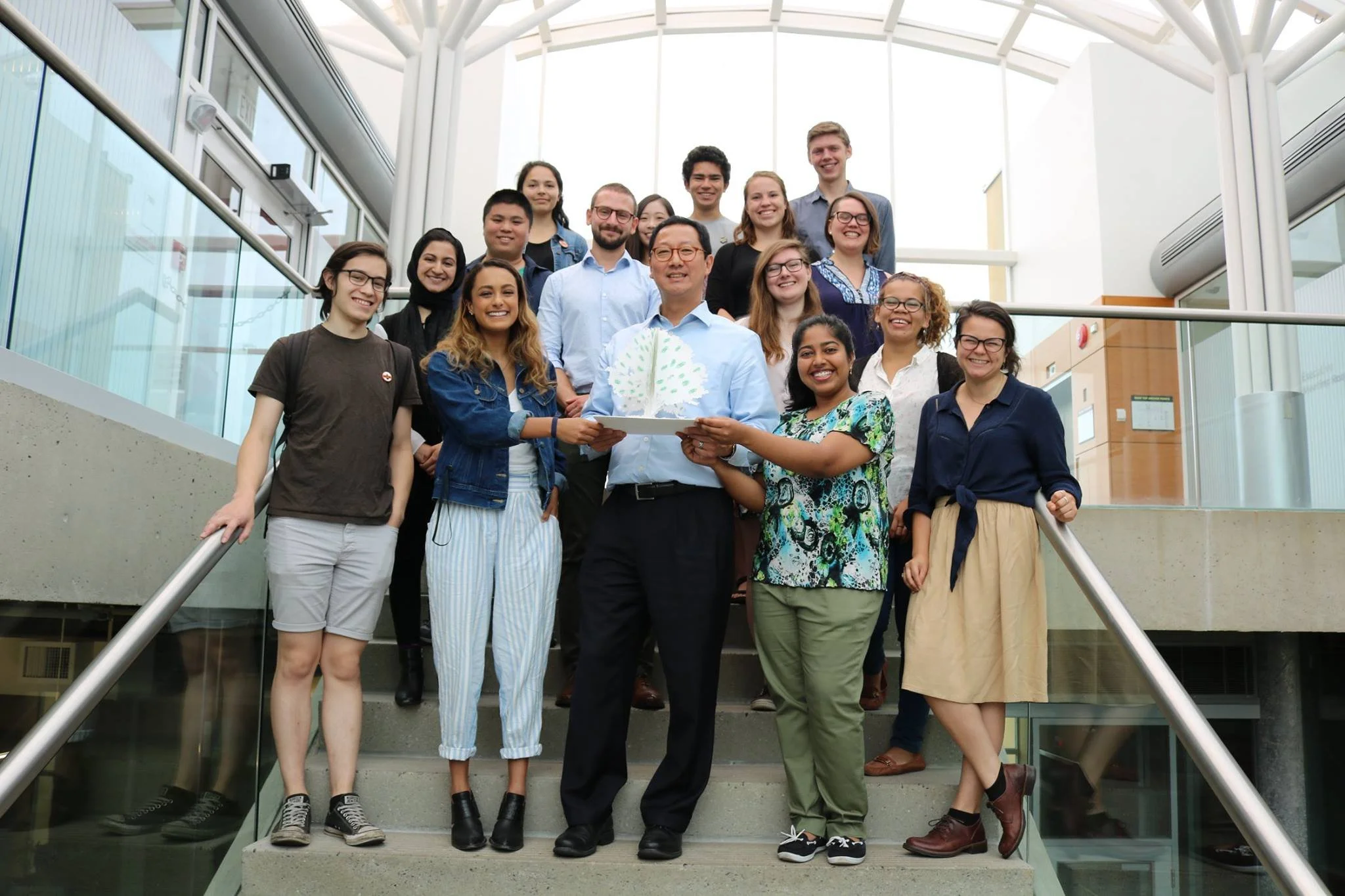 Group of diverse young people and a man holding a decorated cake, standing on a staircase inside a modern building with glass walls and a high ceiling, celebrating an event.