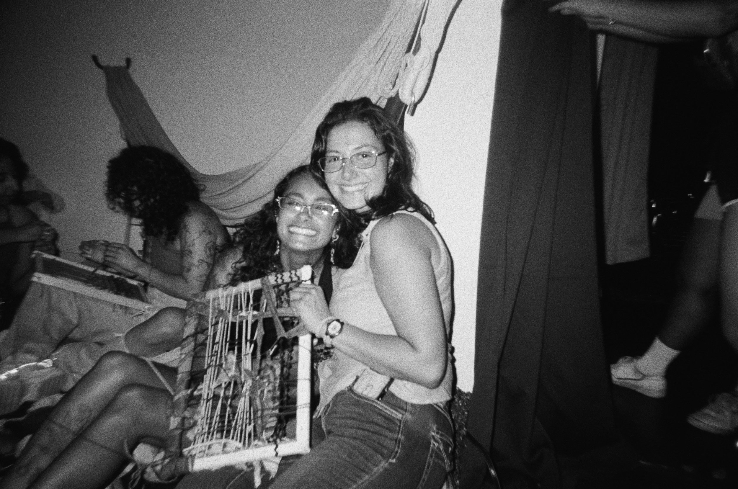 Two young women smiling and embracing each other at a party. One is sitting with a cage on her lap, while the other is standing beside her. Other people are in the background.