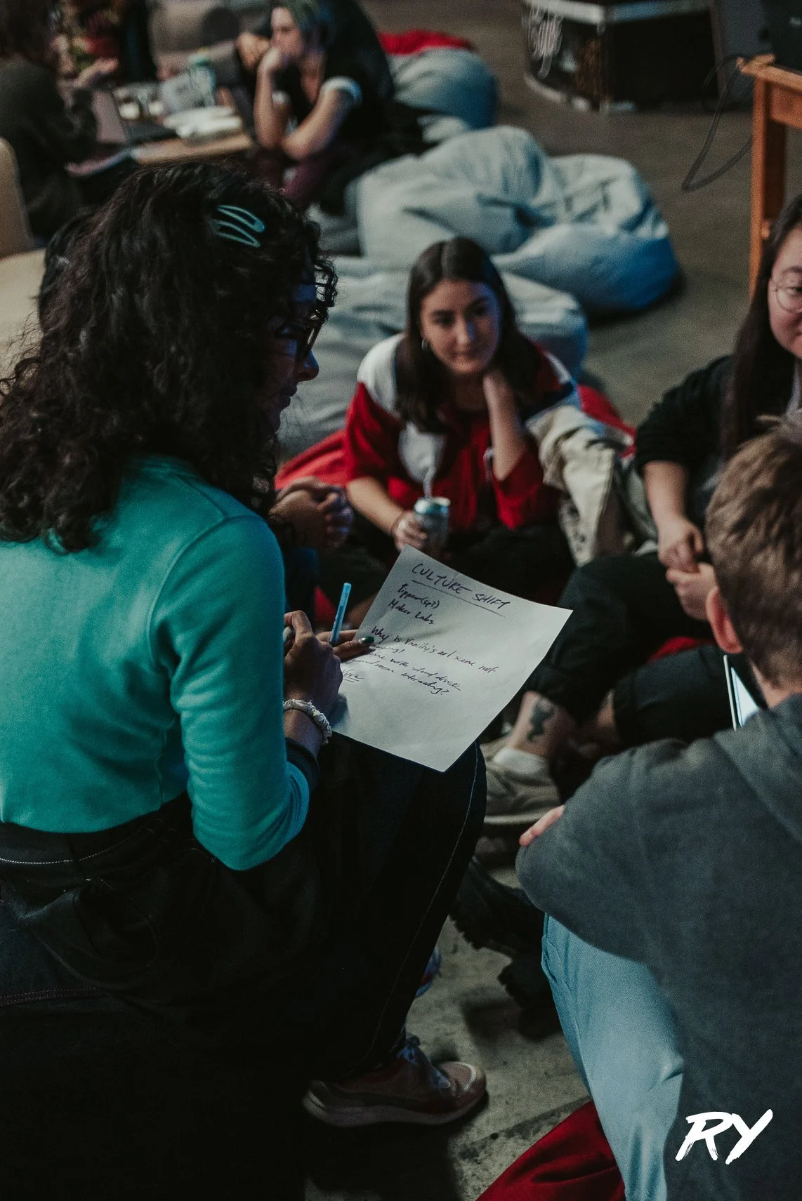 A group of people sitting on the floor and bean bags, engaged in a discussion. A woman with curly dark hair and glasses is holding a notebook and a pen. The notebook has handwritten notes on it. The background shows more people sitting and talking in