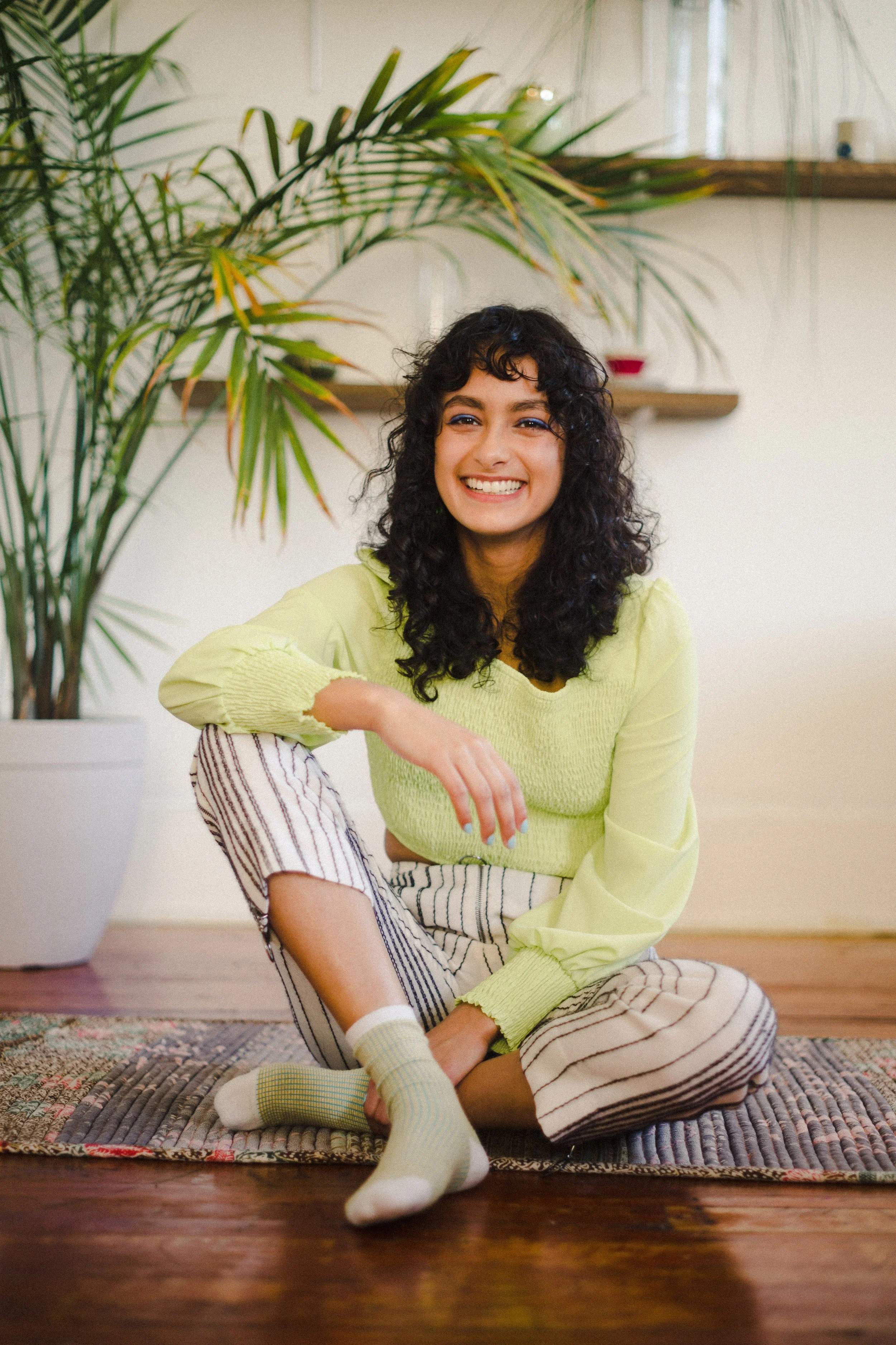 A woman sitting cross-legged on a patterned rug in a bright room, smiling at the camera. She has curly dark hair and is wearing a pale yellow long-sleeve top, striped pajama pants, and matching striped socks. Behind her is a large green plant with lo