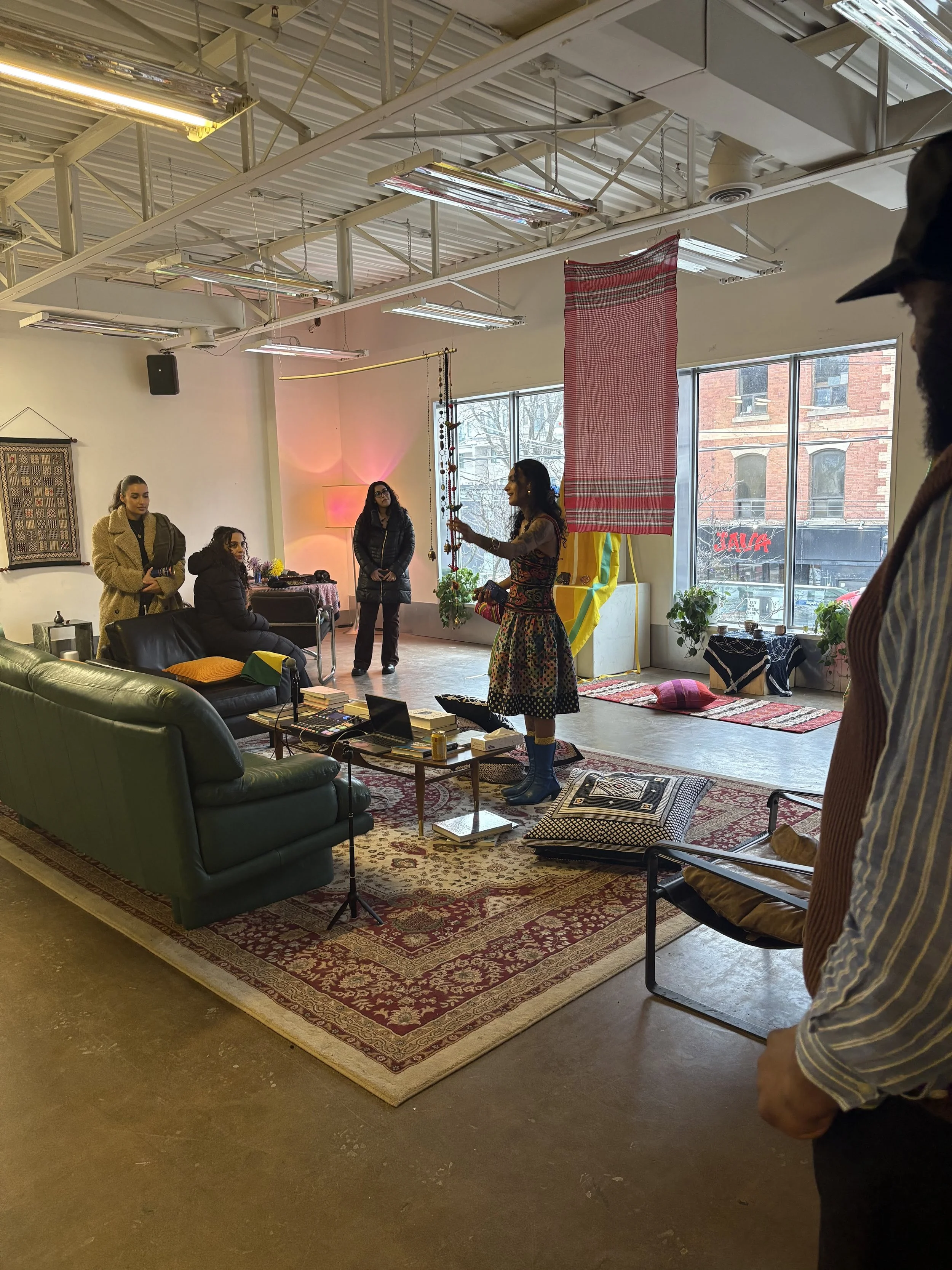 A woman stands in front of a small audience inside a creative studio space with large windows, colorful textiles, and eclectic decor. She appears to be giving a presentation or demonstration, holding a string of objects. The audience is engaged, with
