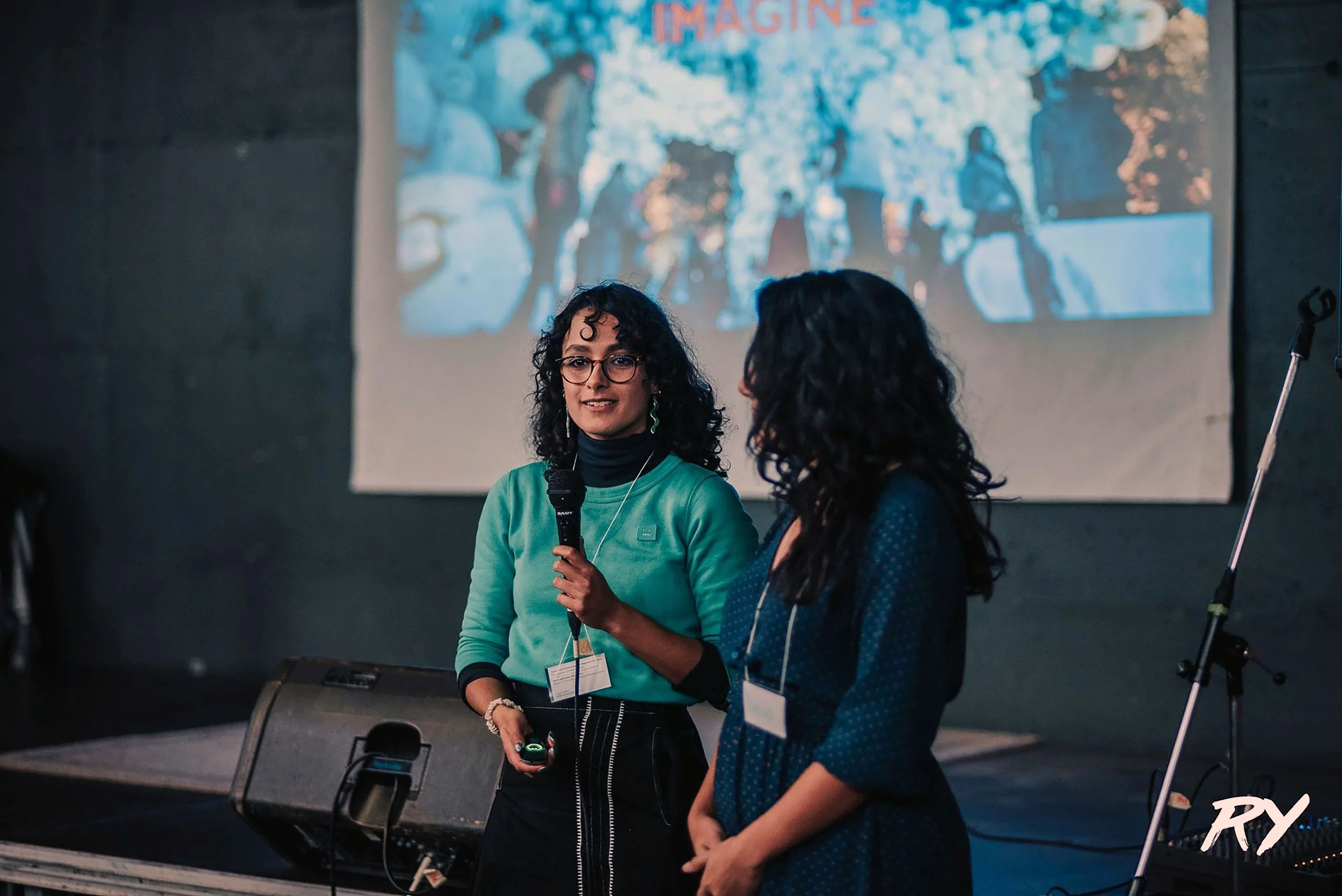 Two women with dark curly hair and glasses are standing on a stage in front of a large screen, one woman is speaking into a microphone while the other listens. The background shows a blurry image of people and trees with the word 'IMAGINE' visible at