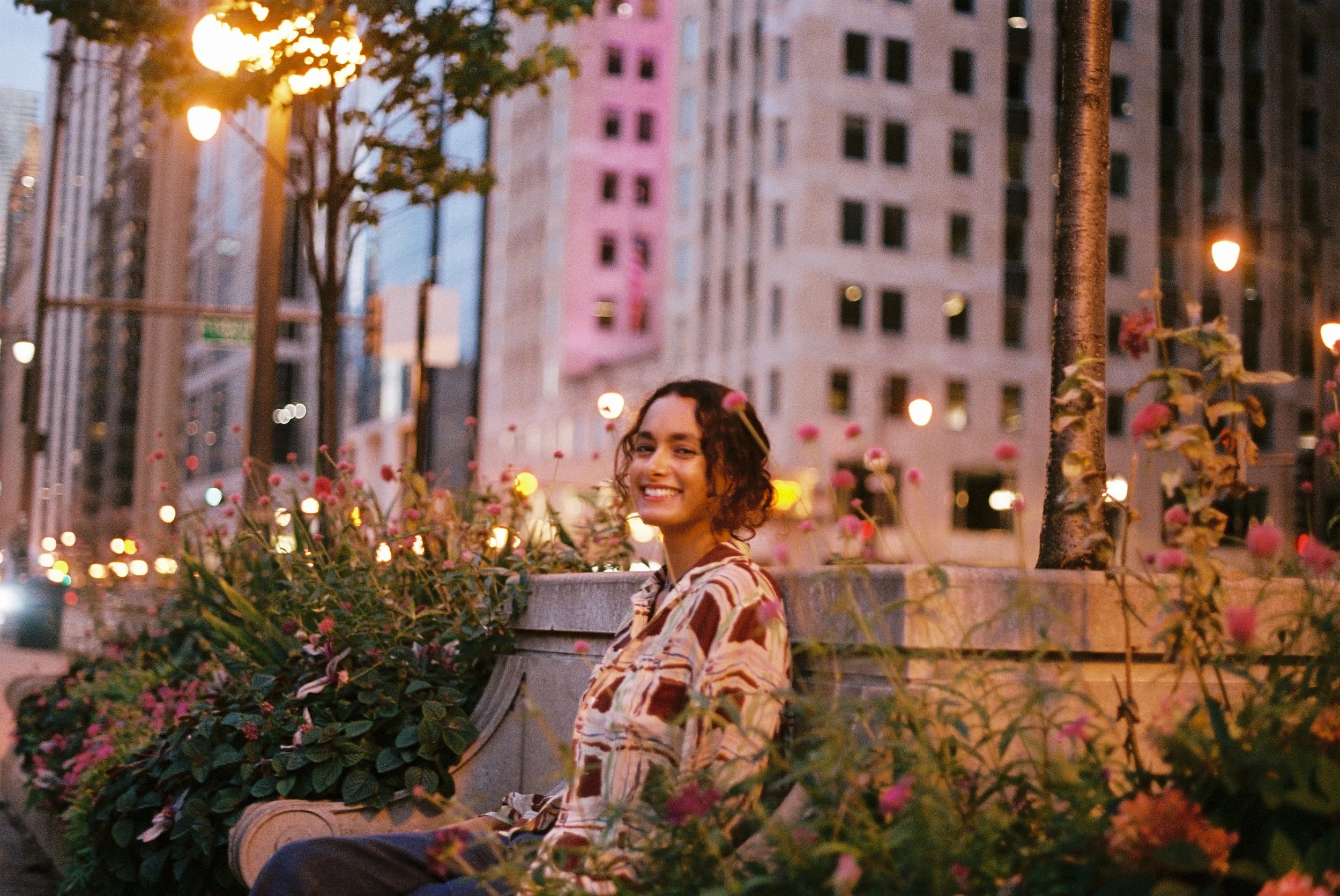 A woman with curly hair smiling while sitting on a bench surrounded by pink flowers in an urban park at sunset.