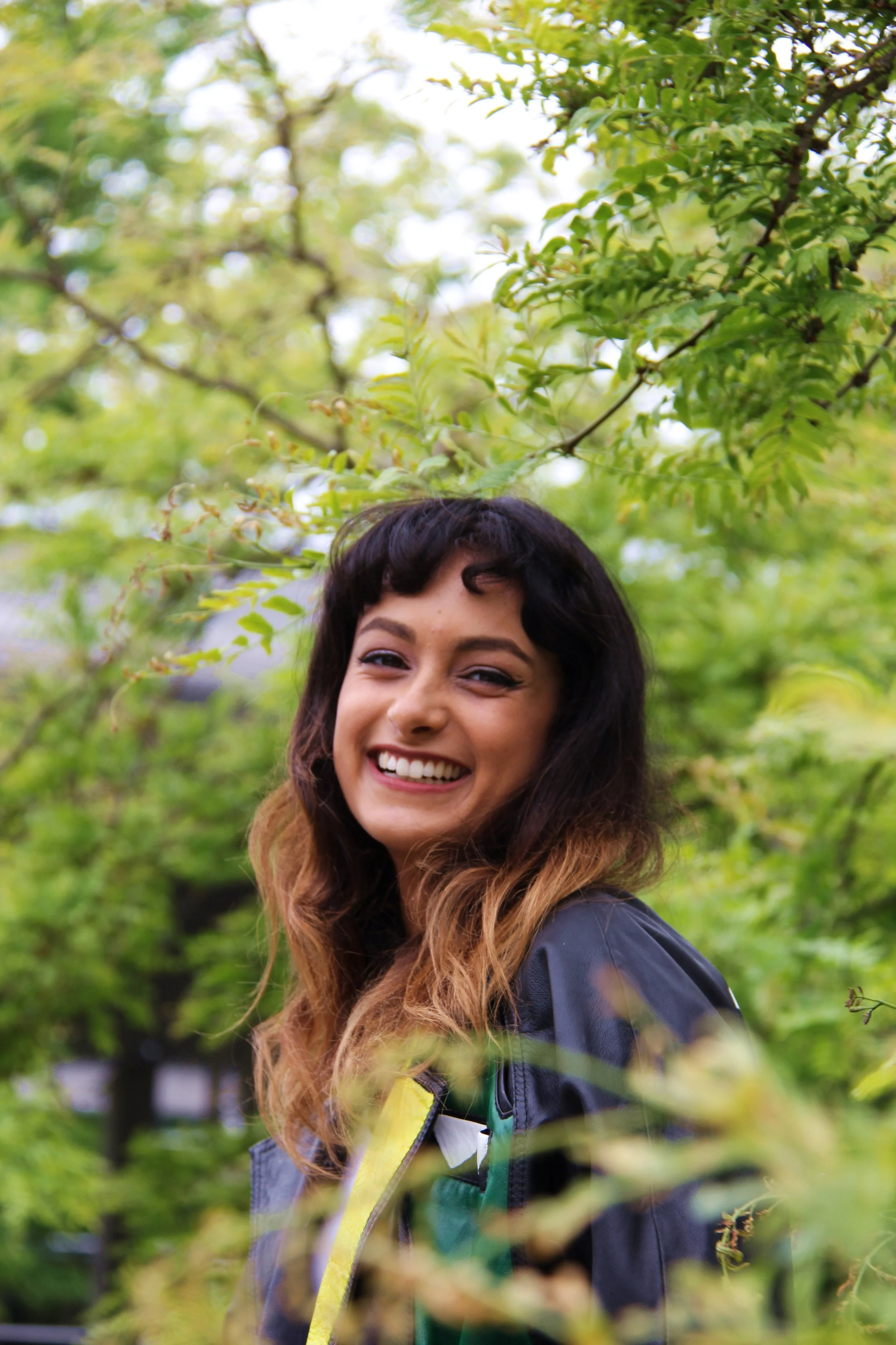 A young woman with wavy brown hair smiling outdoors among green trees, wearing a black jacket with yellow and green accents.