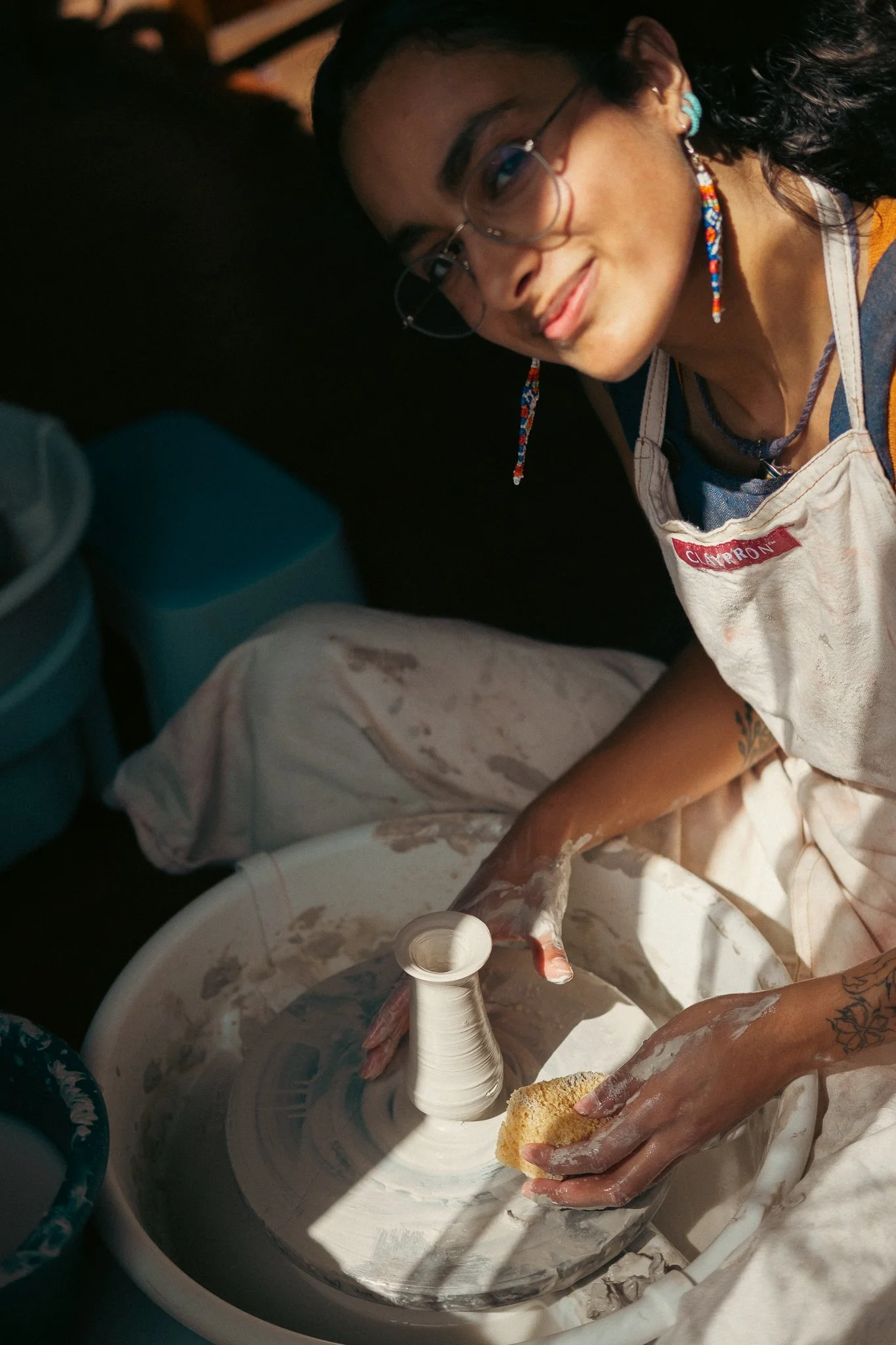 A woman with glasses and colorful earrings is pottery making on a spinning wheel, shaping clay with her hands, wearing a beige apron that says 'Clément' and using a sponge.