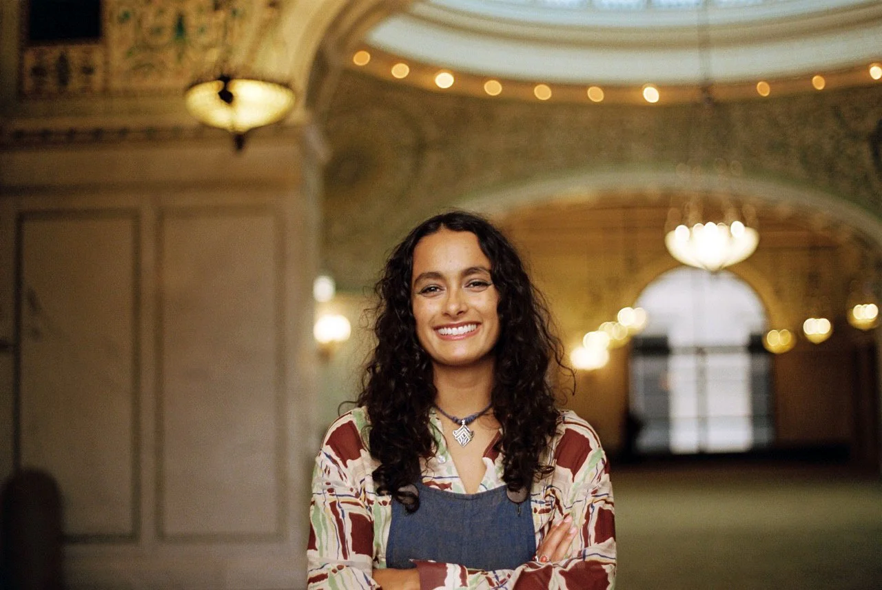 A young woman with curly dark hair, smiling with cross arms, stands in a grand, ornate hallway with chandeliers and decorative lighting.