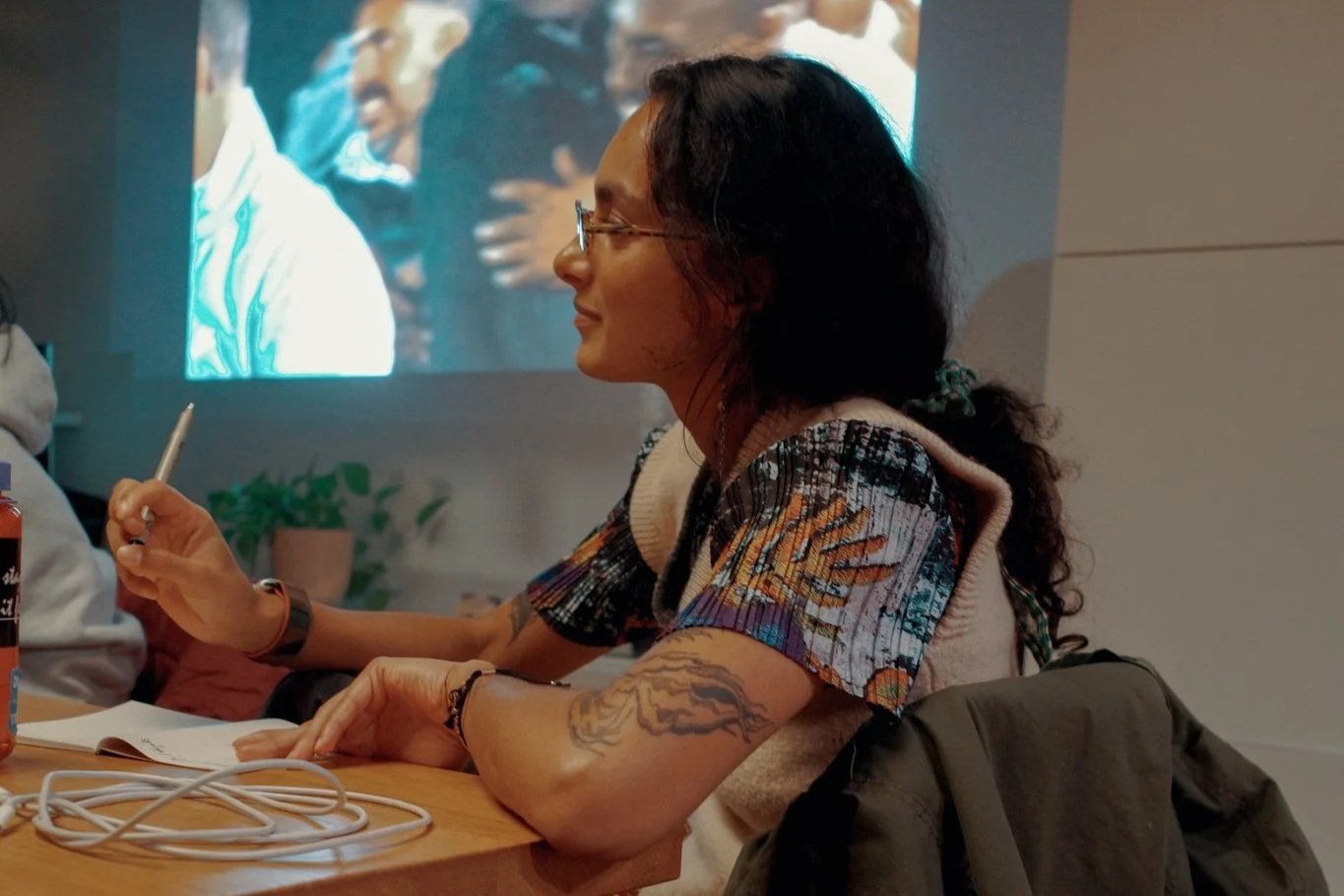 Woman with glasses and tattoos sitting at a table with a notebook, pen, and cables, smiling and engaged in a discussion while a video is projected on the wall behind her.