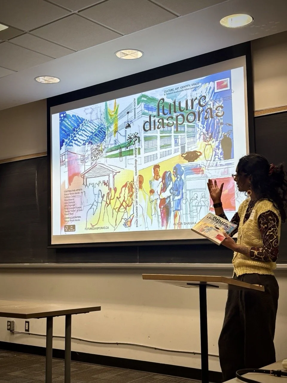 A woman standing in front of a projection screen giving a presentation about future diaspora, culture, and art. The screen displays an illustrated poster with people, buildings, and colorful artwork.