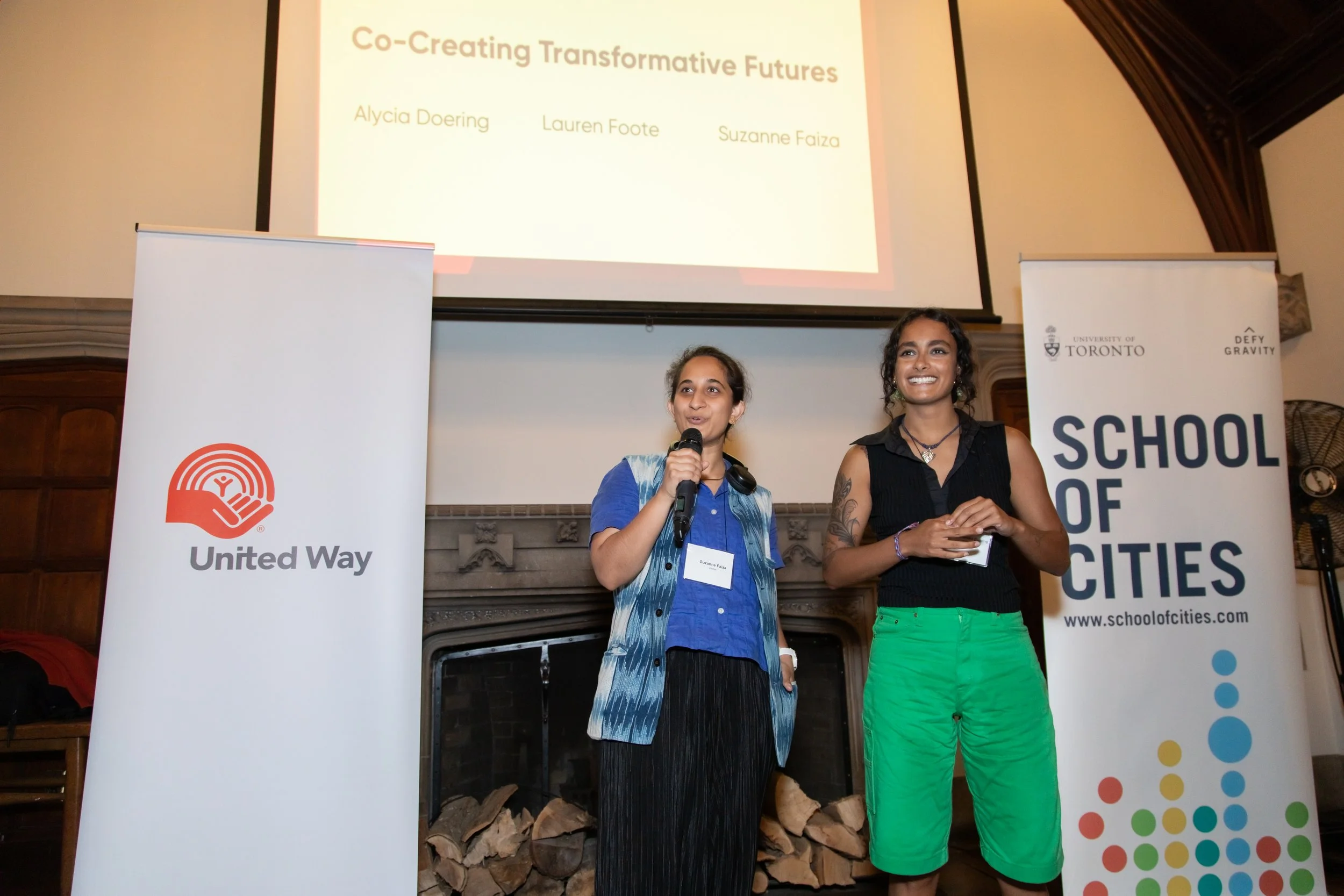 Two women speaking at a podium during a presentation with banners for United Way and School of Cities in the background.