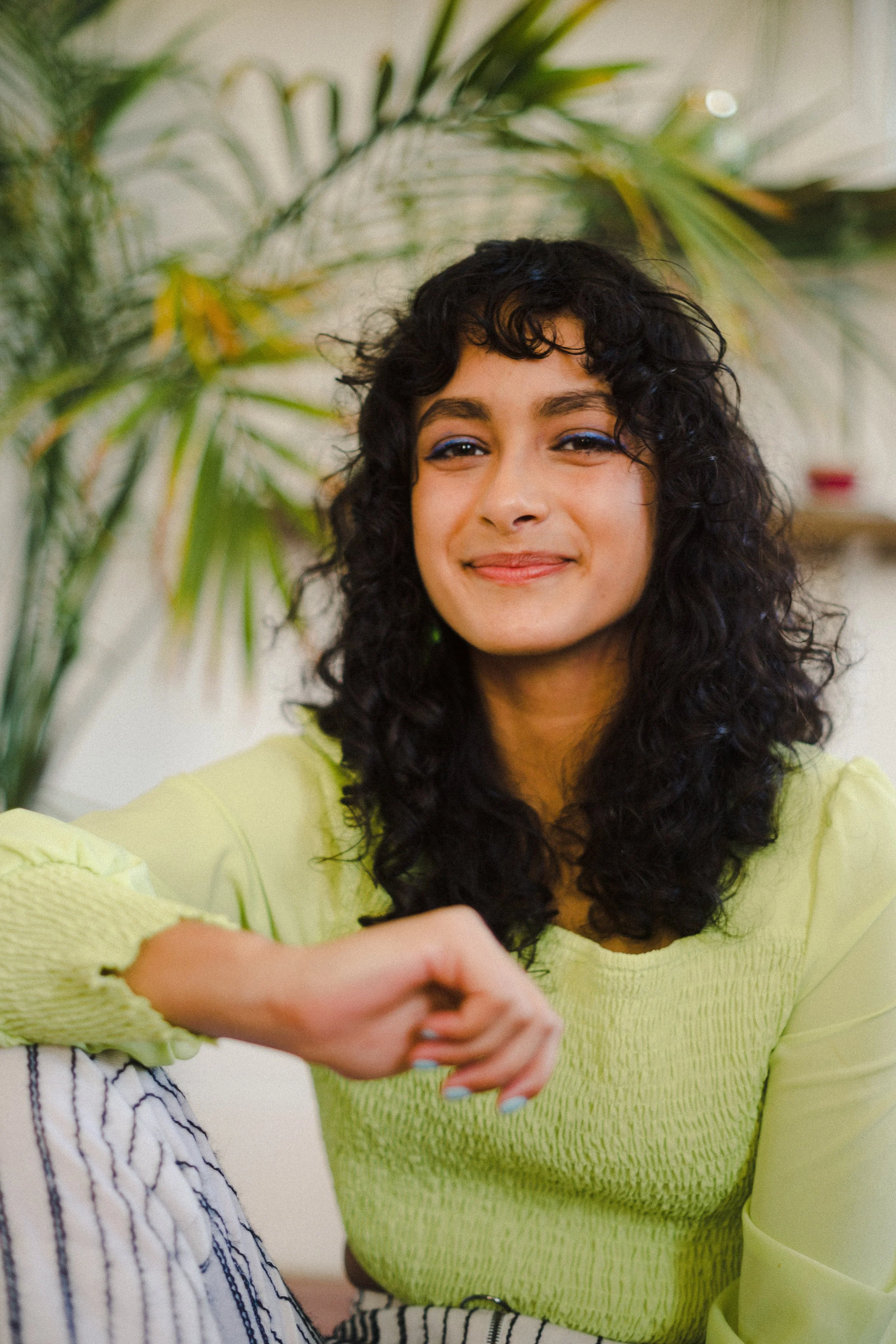 A woman with curly dark hair, wearing a light green top, smiling in a room with a plant in the background.