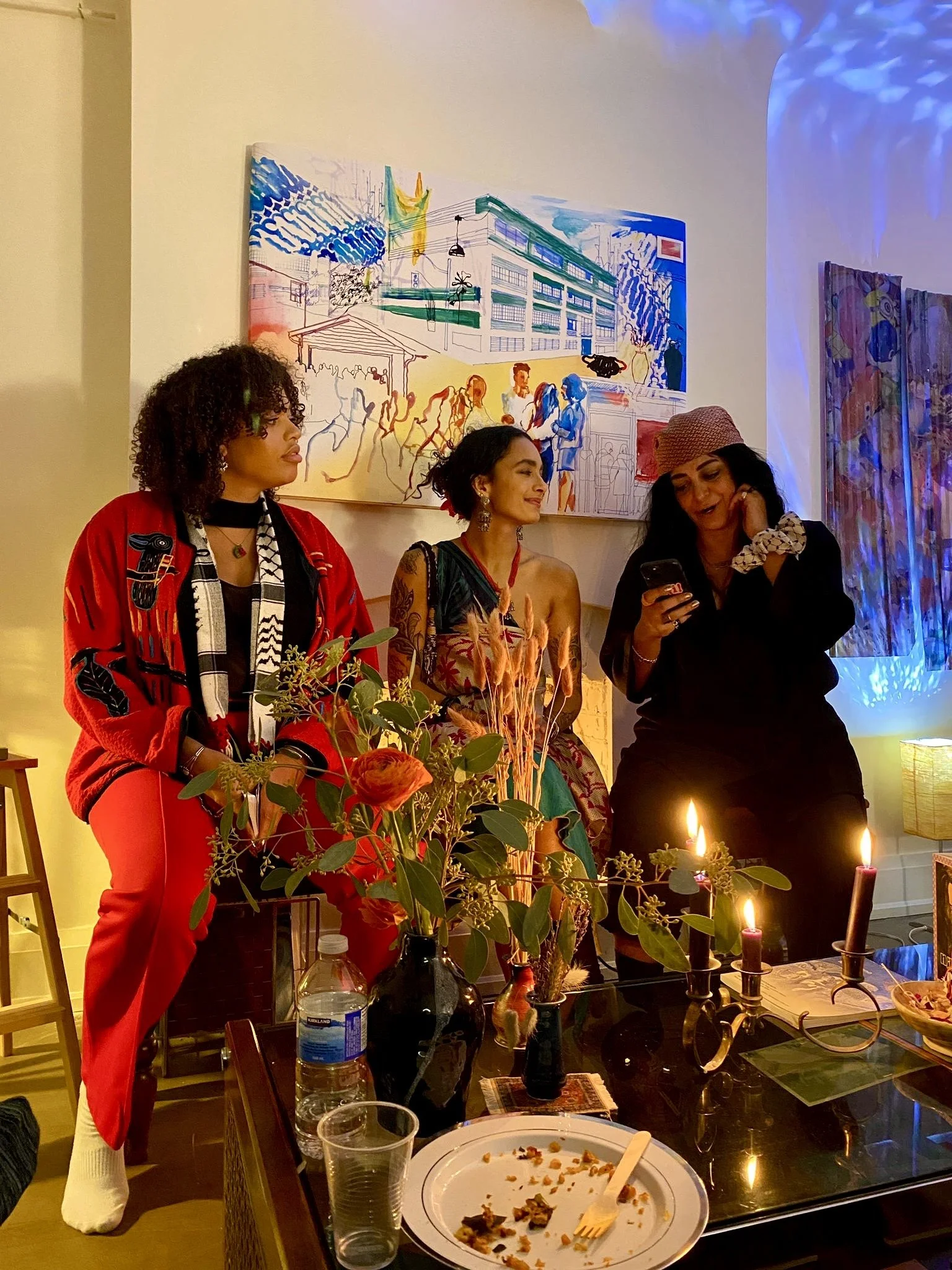 Three women sitting at a table, one reading a phone, with a colorful painting in the background. The table has a vase with flowers, candles, a water bottle, and a used plate.