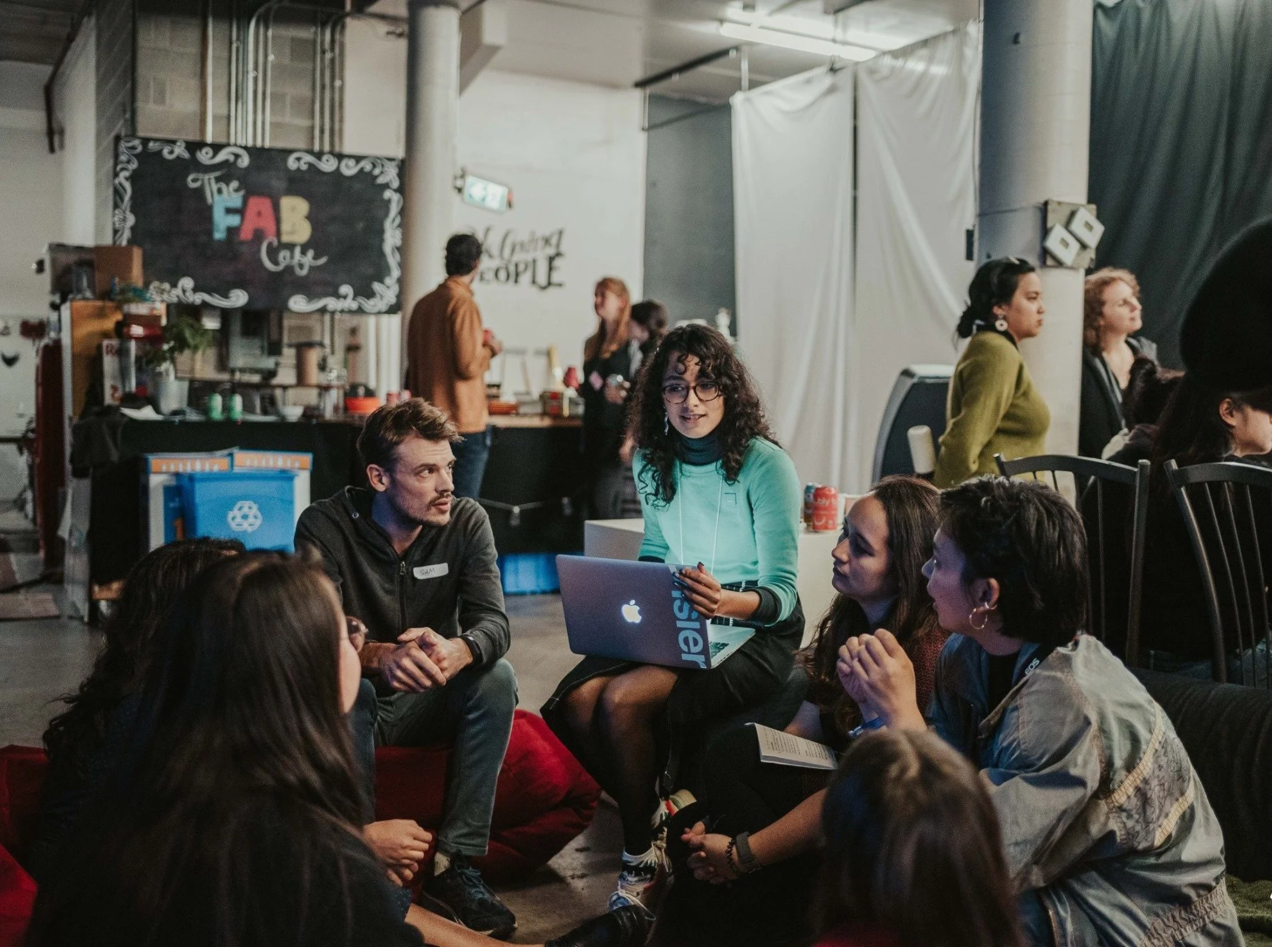 Group of young adults sitting on the floor and engaging in conversation at a casual social event in a large indoor space with a food stand and decorative wall art in the background.