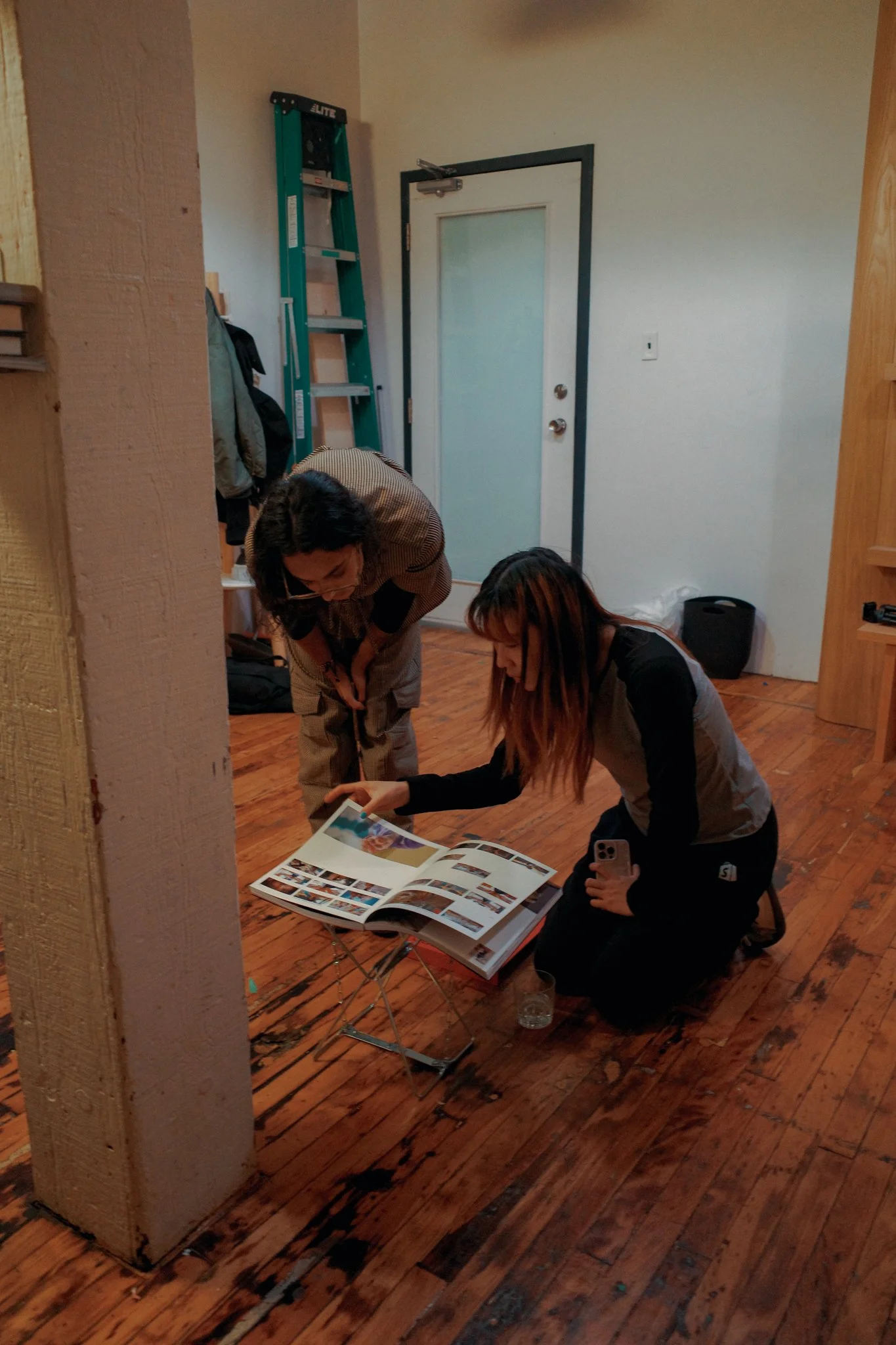 Two women are looking at a photo album on a small table inside a room with wooden flooring. One woman is kneeling while the other is standing, both focused on the album. A ladder is leaning against the wall near a door.