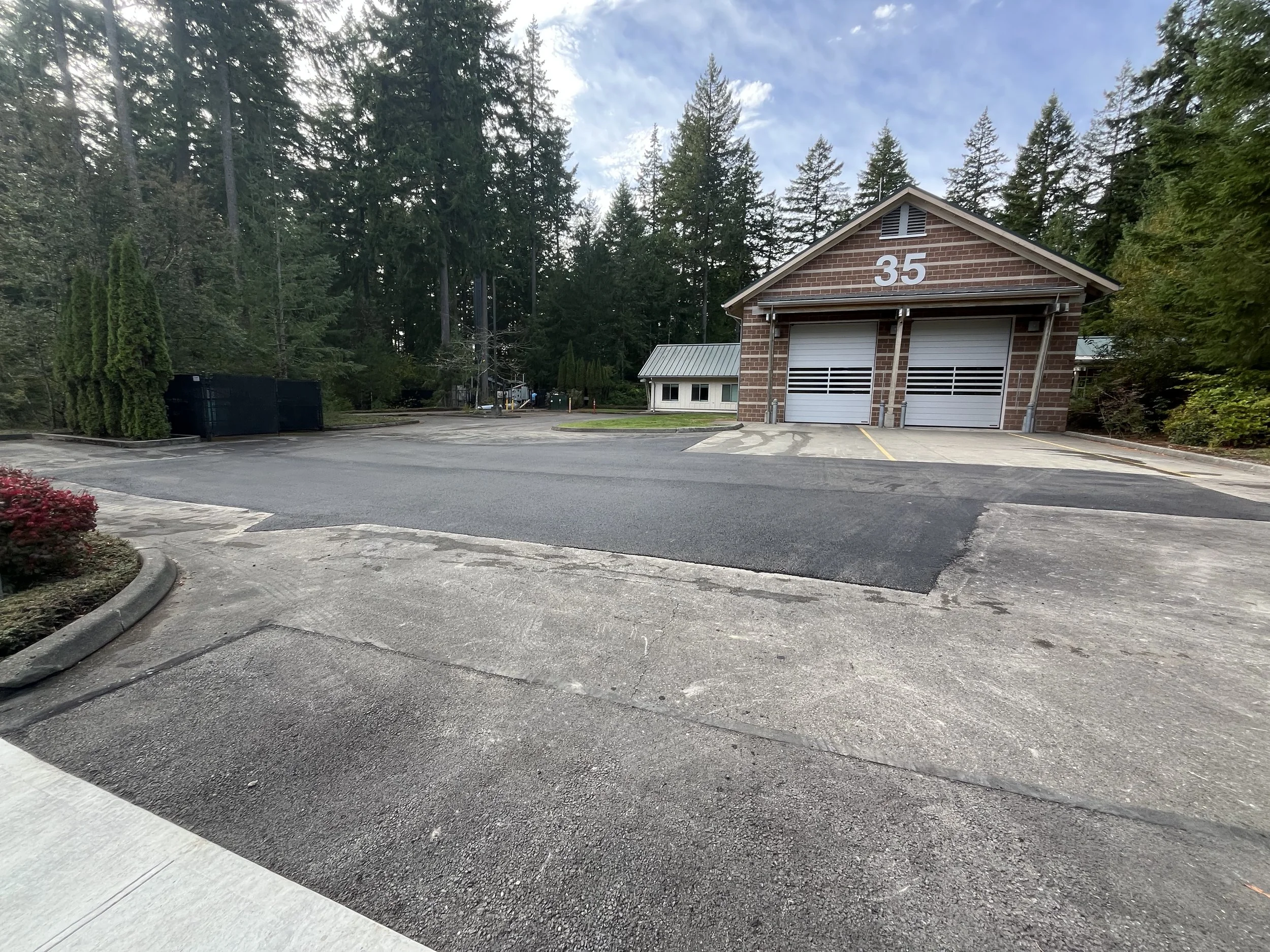 A parking lot in front of a small brick building with two garage doors numbered 35, surrounded by trees and a partly cloudy sky.