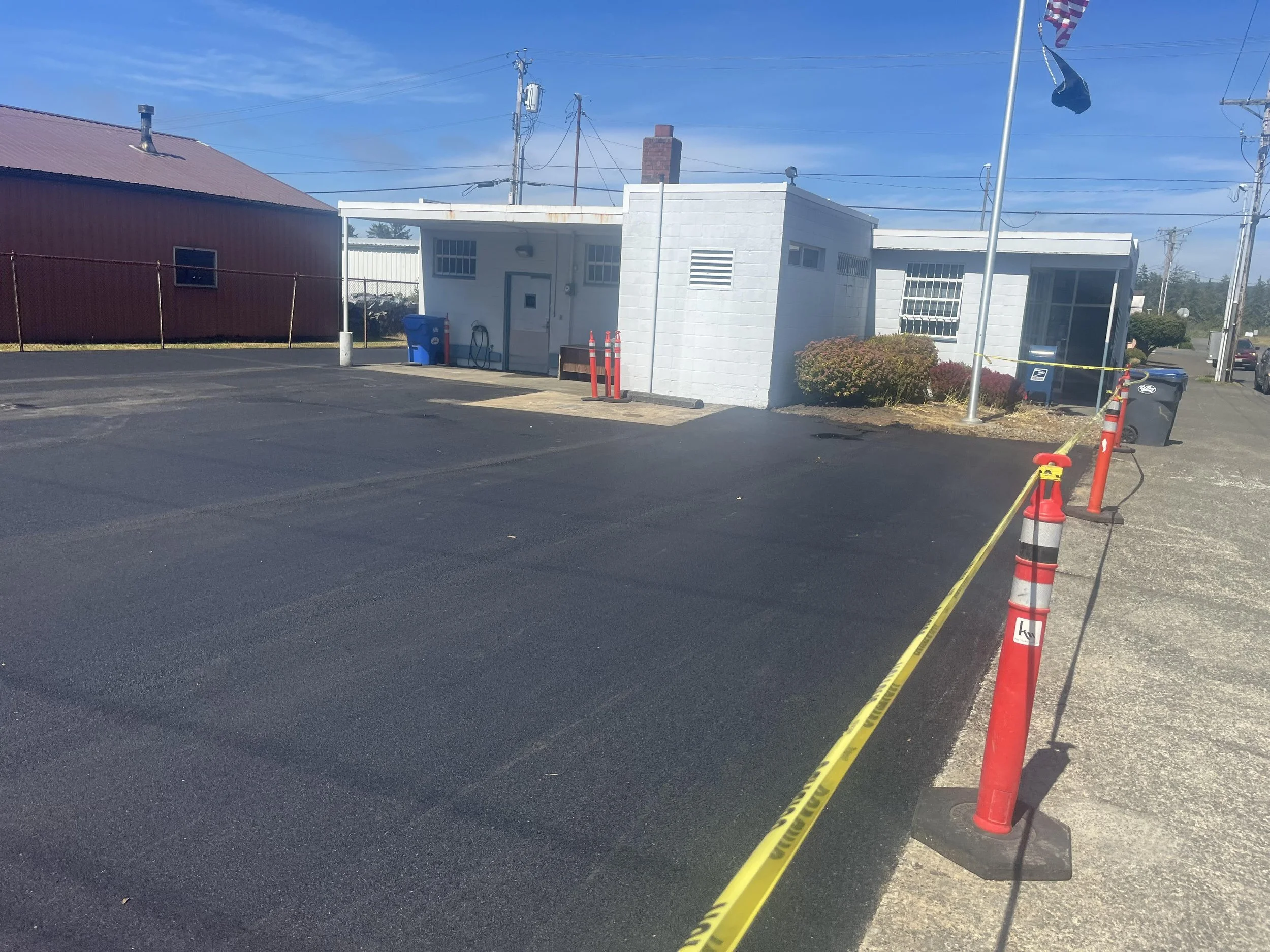 A freshly paved parking lot with black asphalt, surrounded by orange and yellow safety cones and yellow caution tape, outside a small white building under a blue sky.