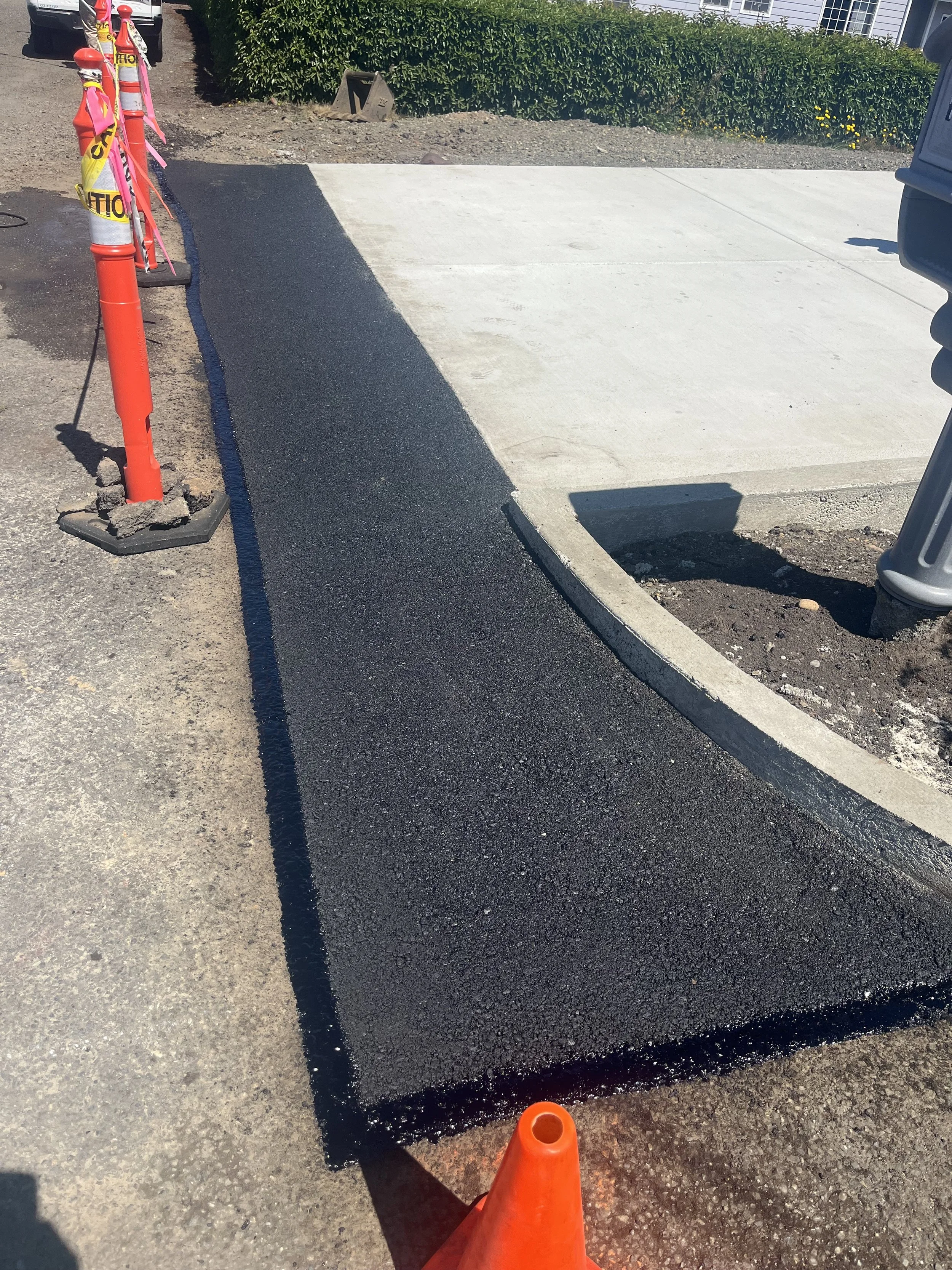 Freshly paved black asphalt sidewalk alongside concrete curb and driveway, with construction cones and warning flags around.