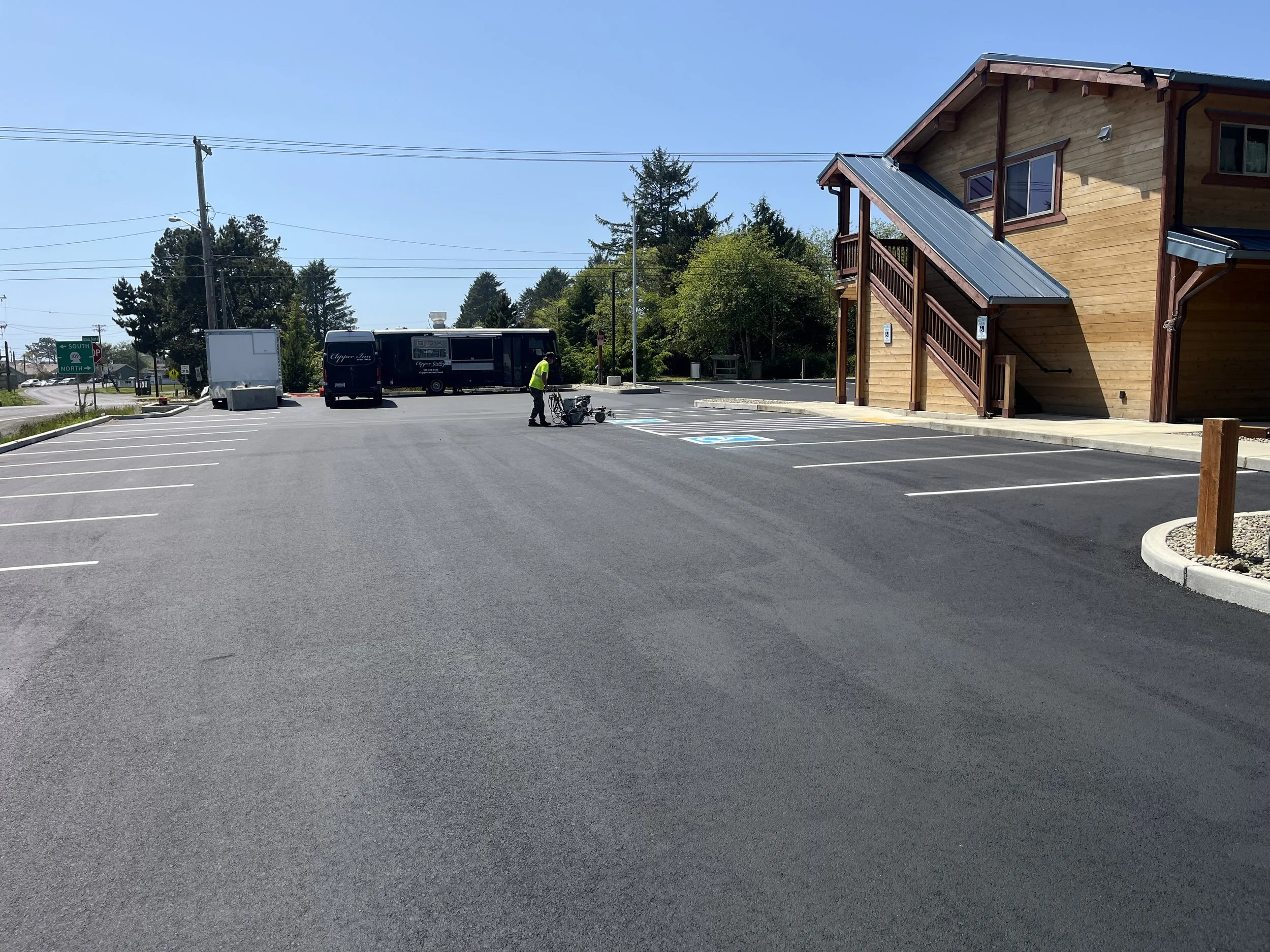 A parking lot with freshly paved asphalt, a handicapped parking space, a person in a high-visibility vest working, a bus, and a building with a wooden exterior and stairway.