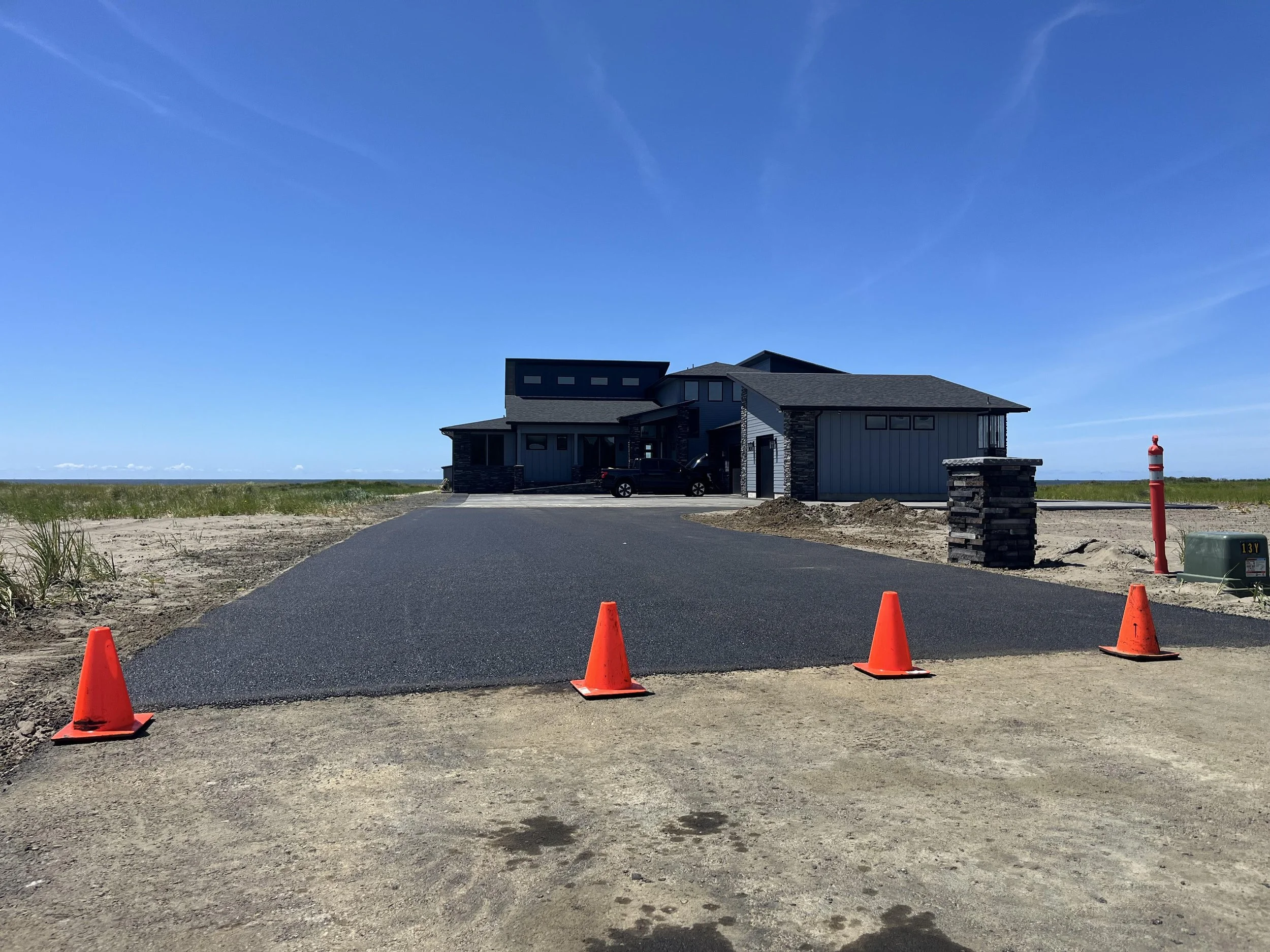 A modern house under construction with a freshly paved driveway blocked by orange traffic cones on a partly cloudy day with a bright blue sky.