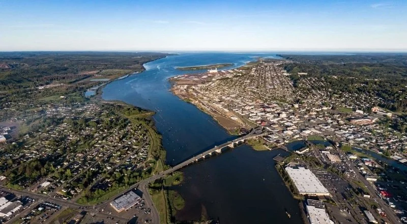 An aerial view of a wide river flowing through an urban area with bridges, residential neighborhoods, commercial buildings, and green spaces on either side under a clear blue sky.
