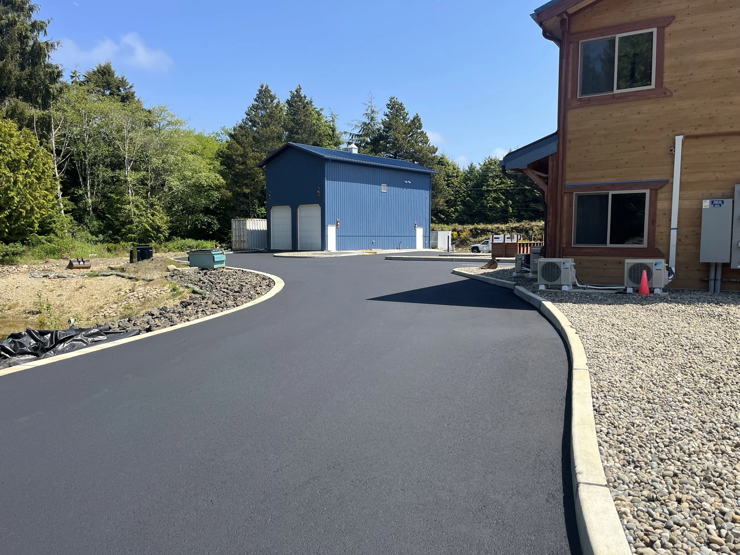 Newly paved black asphalt driveway winding past two buildings, one wooden with visible windows and air conditioning units, and a blue metal building in the background, with trees and a clear blue sky.