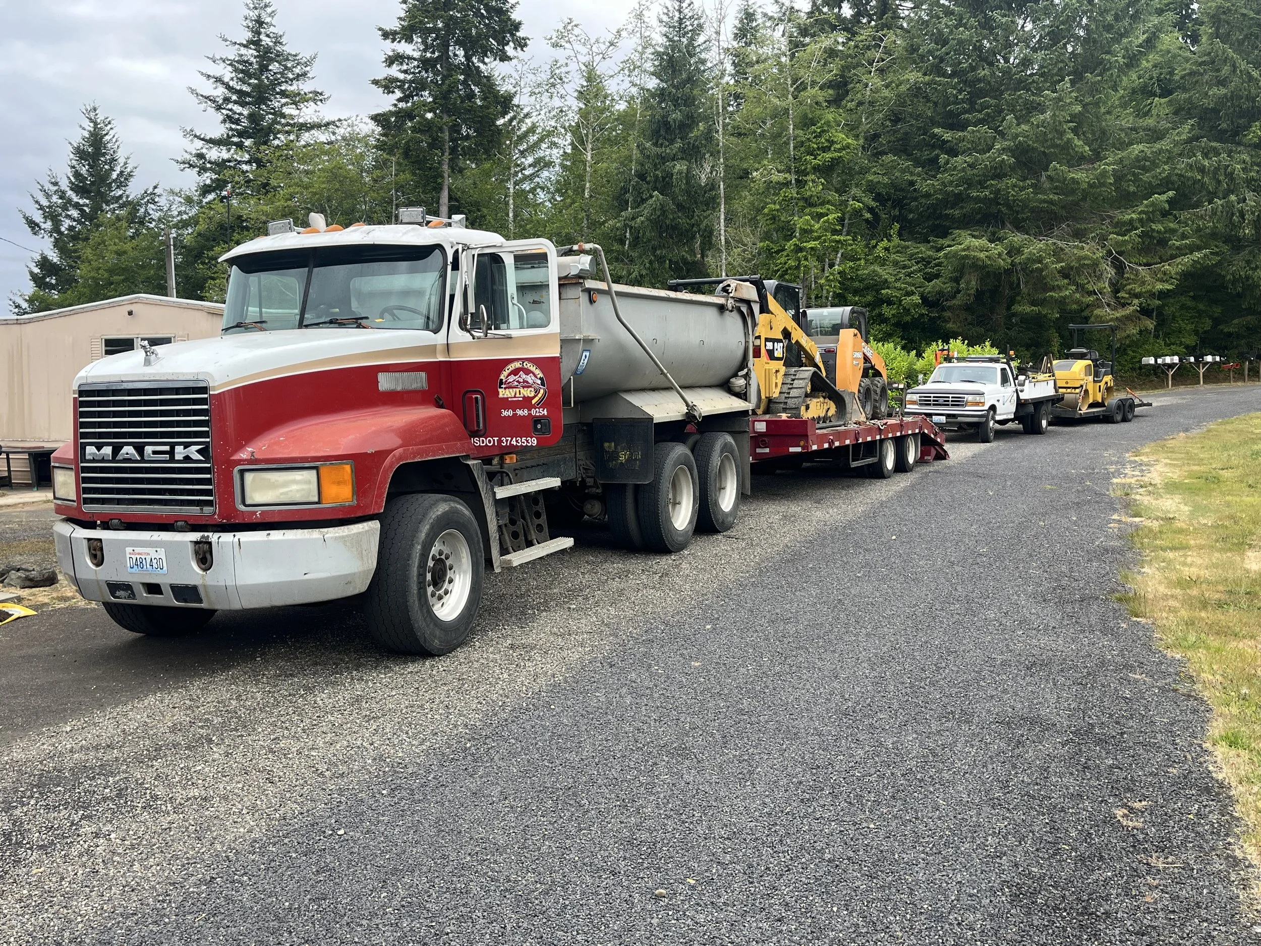 A large Mack truck with a flatbed trailer carrying construction vehicles and equipment, parked on a gravel driveway with trees in the background.