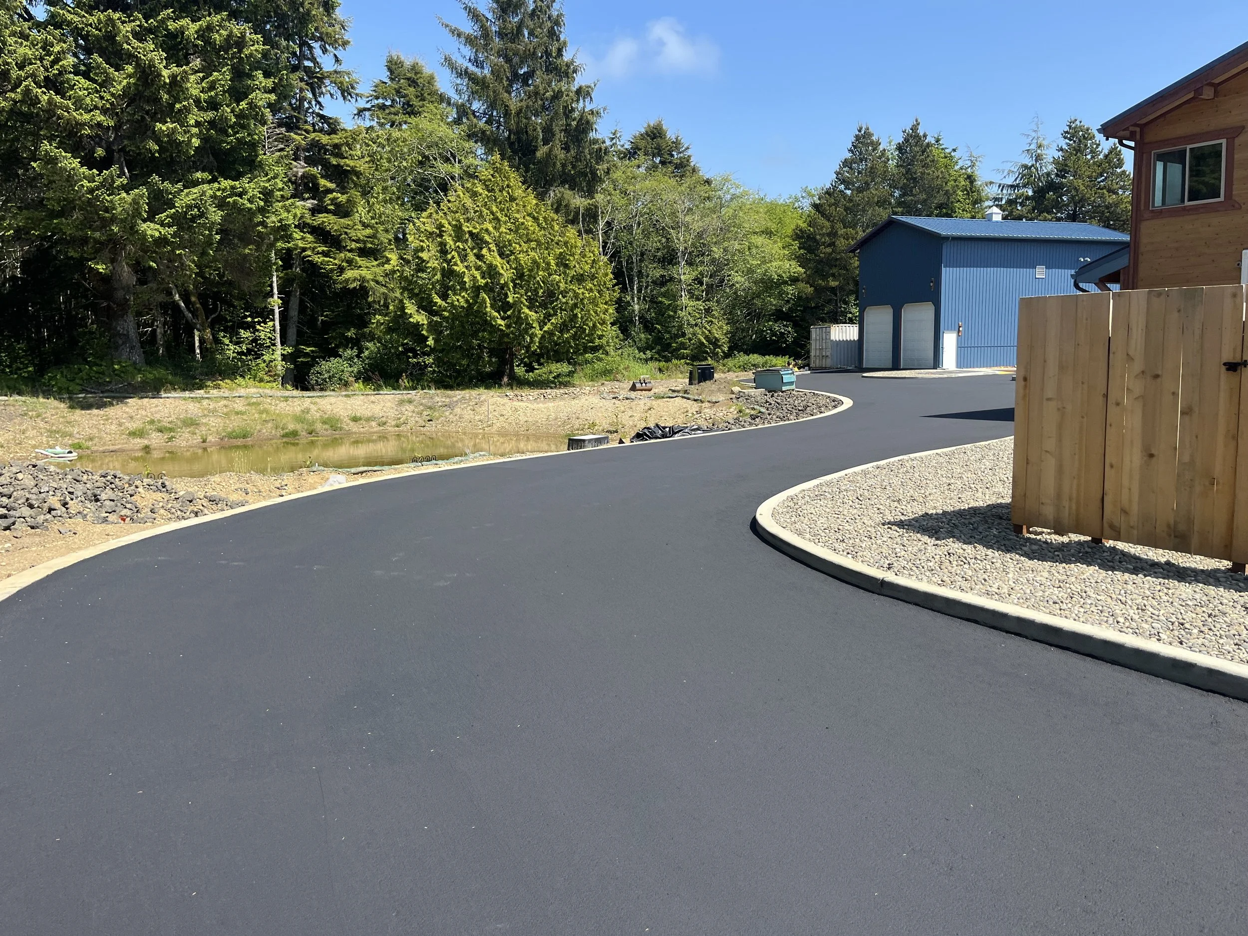 Curved newly paved blacktop driveway next to a wooden fence, with a multicolored house and a blue shed in a wooded area on a sunny day.