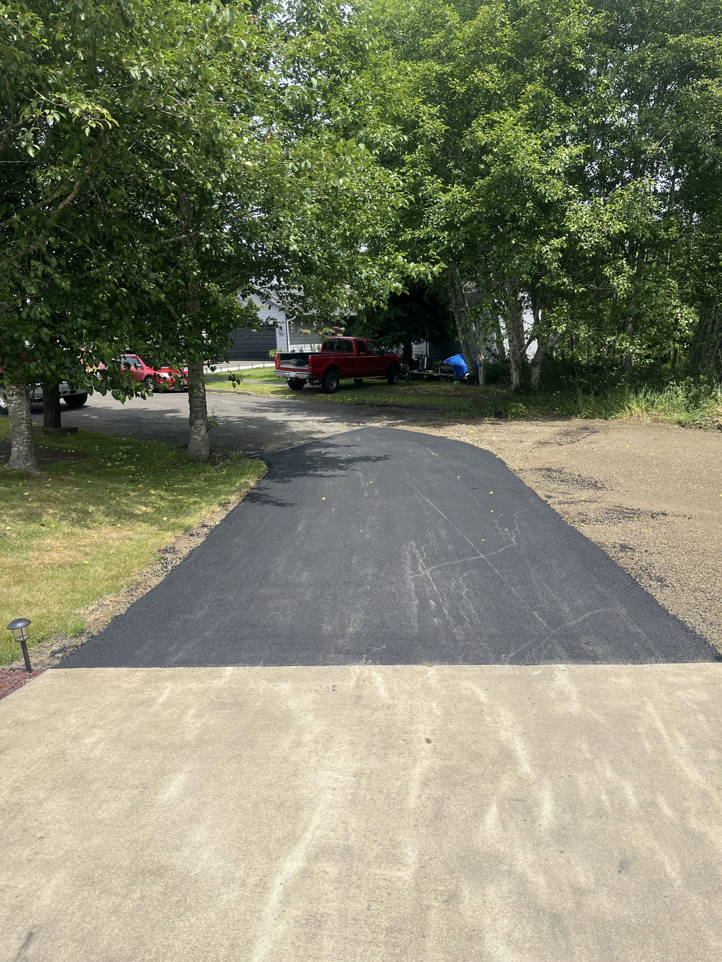A freshly paved black asphalt driveway leading from concrete to a grassy yard, with trees in the background and a parking area with vehicles including a red truck.