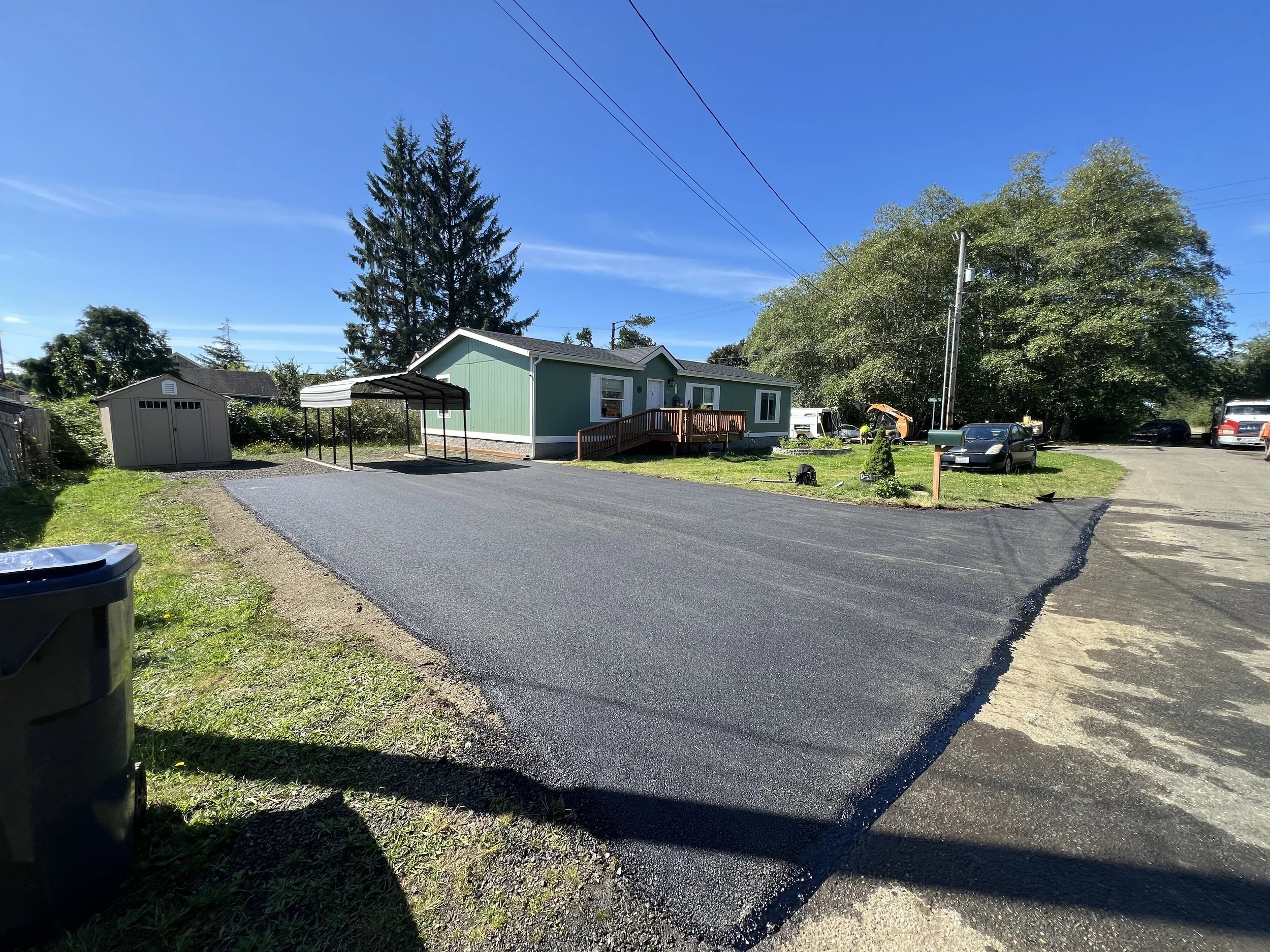 A newly paved black asphalt driveway in front of a green house with a small wooden ramp, a shed on the left, and a yellow excavator and several parked cars in the background on a bright sunny day.