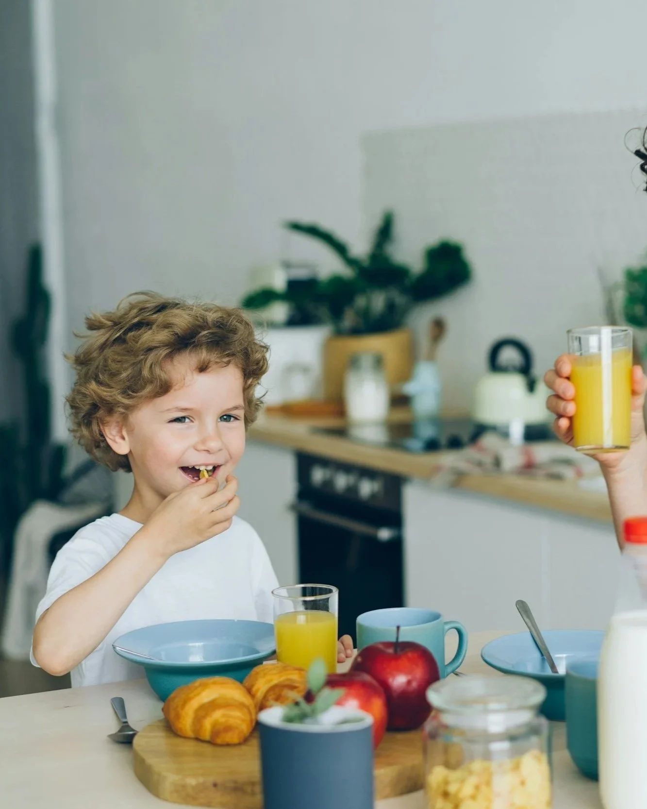 A young boy with curly hair enjoying breakfast, with a glass of orange juice in front of him. The table is set with breakfast items, including apples, croissants, a mug, and a bowl. Someone is pouring more orange juice into a glass.