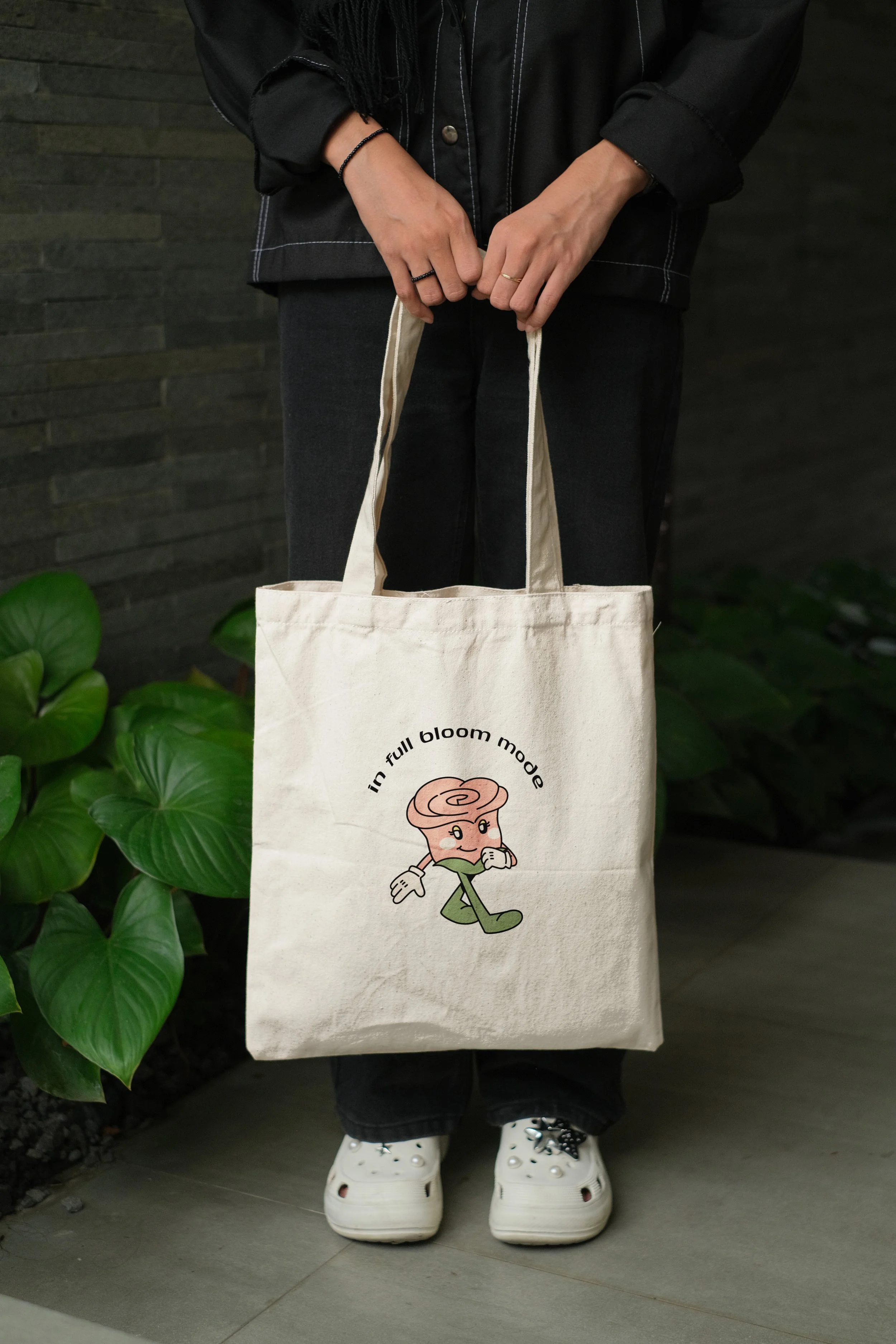 Person holding a beige tote bag with the phrase "in full bloom mode" and a cartoon character of a pink rose with a face, arms, and legs, walking and smiling, standing on a gray outdoor surface with green plants and a dark gray wall in the background.