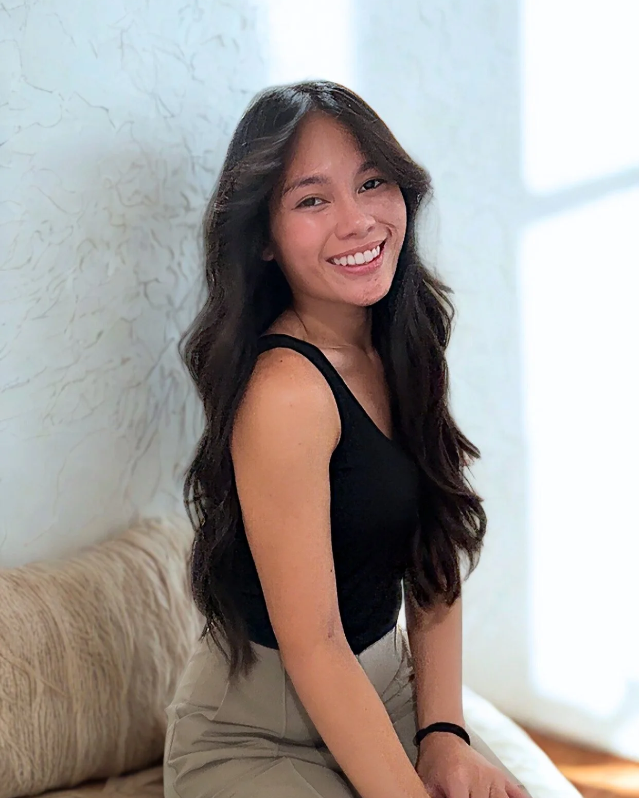 A young woman with long, dark wavy hair, smiling and looking at the camera, wearing a black sleeveless top and light-colored pants, sitting on a beige couch near a textured white wall.