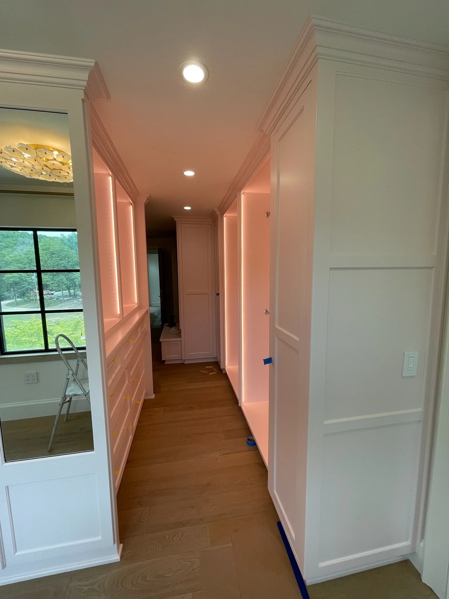 A hallway in a house under construction with white cabinets on both sides and built-in lighting, hardwood floors, and a large window showing a green outdoor scene.