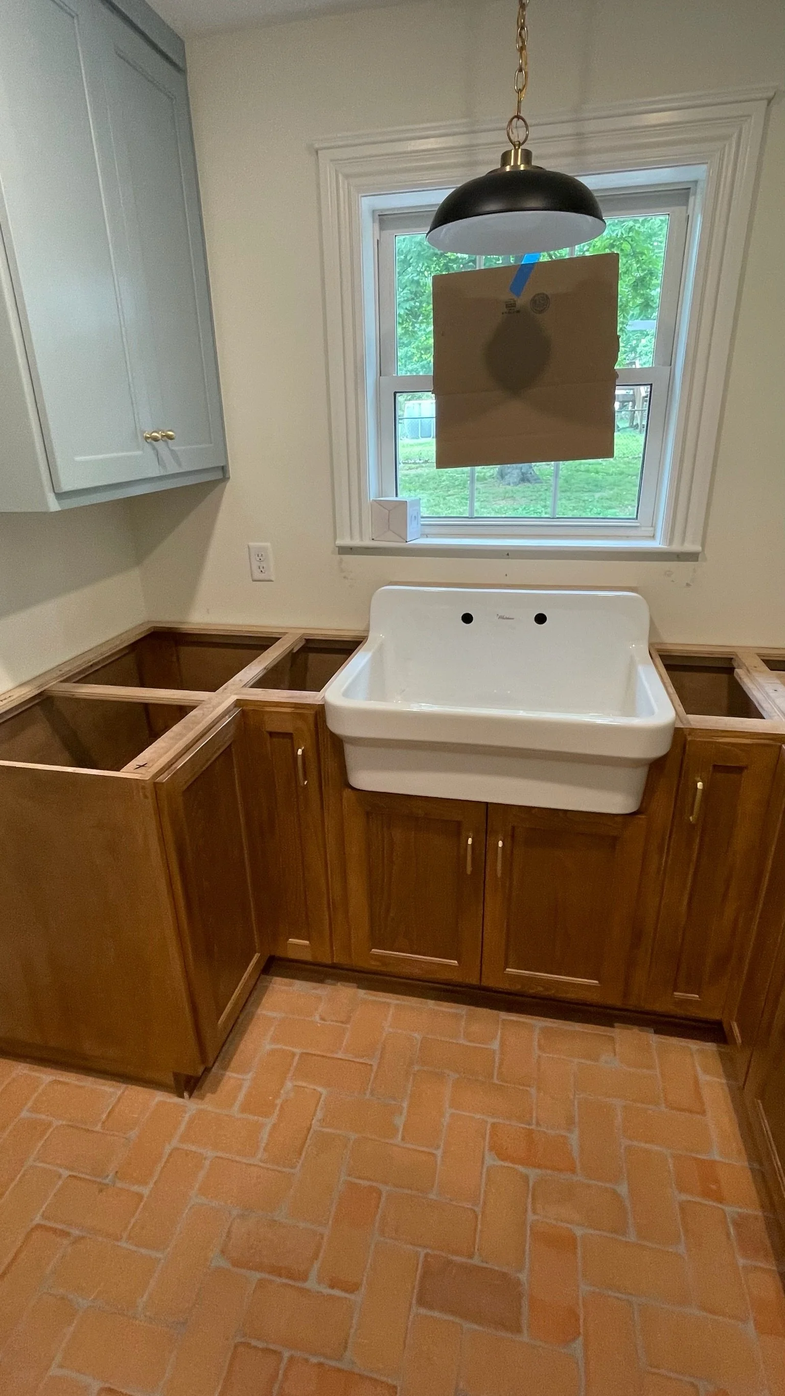 Kitchen with wooden cabinets, brick tile floor, and a white sink underneath a window with a piece of cardboard taped in front of the glass. Overhead light fixture hanging from the ceiling.