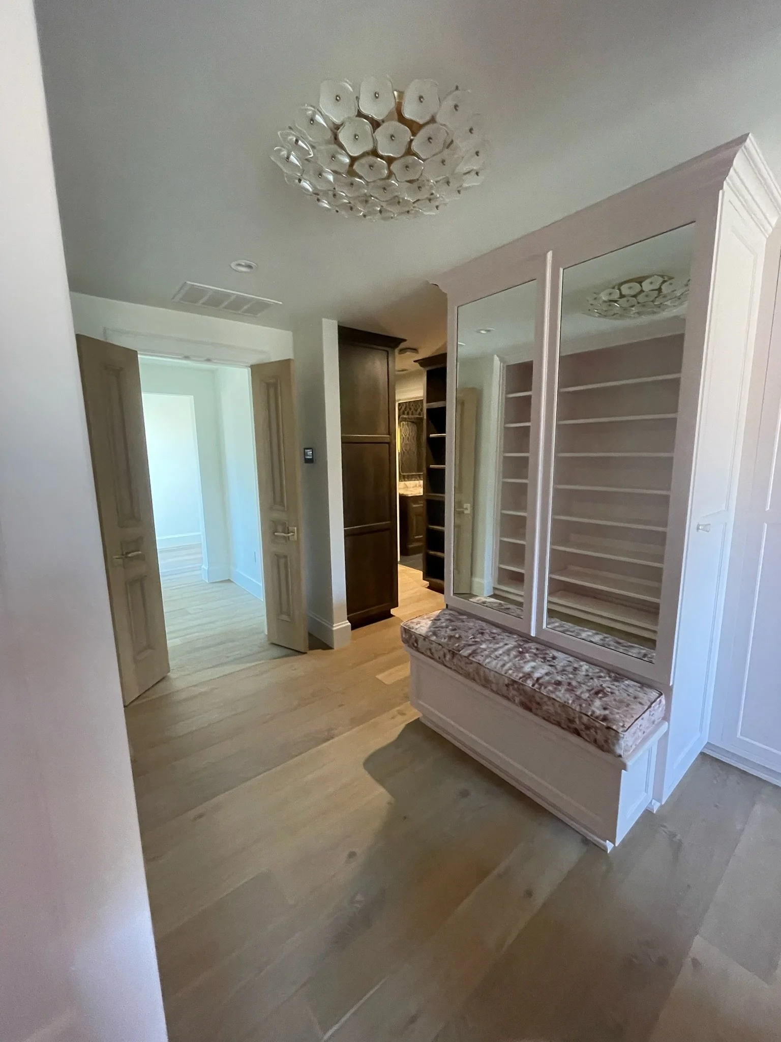A hallway with wooden floors, a white built-in cabinet with mirror doors and a cushioned bench, open doors leading to other rooms, and a decorative ceiling light fixture.