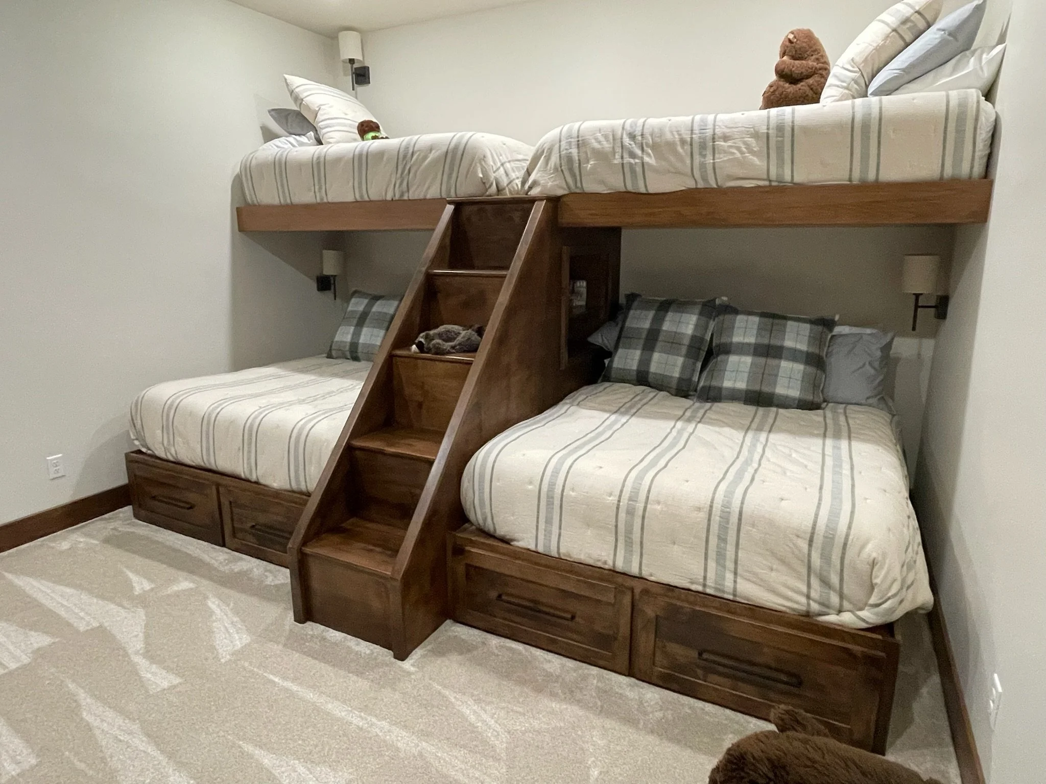 A wooden bunk bed with two full-sized beds, a ladder in the middle, and storage drawers underneath each bed. The beds have cream-colored plaid bedding and pillows, with plush toys on each bed.