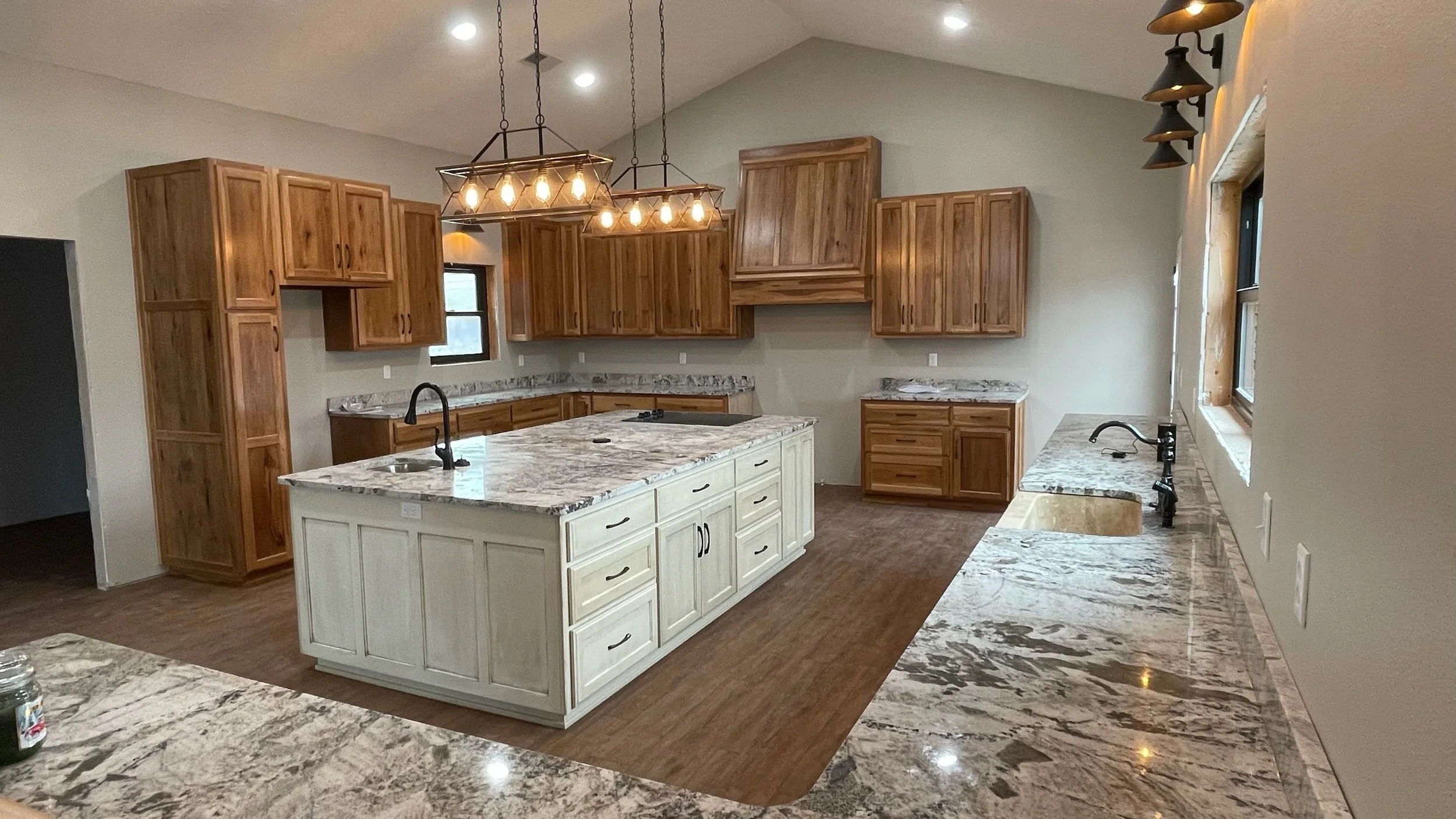 Modern kitchen with wooden cabinets, granite countertops, a large white central island with drawers, stainless steel sink, black faucet, and a contemporary light fixture hanging above. The kitchen has a wood floor, multiple windows, and under-cabinet